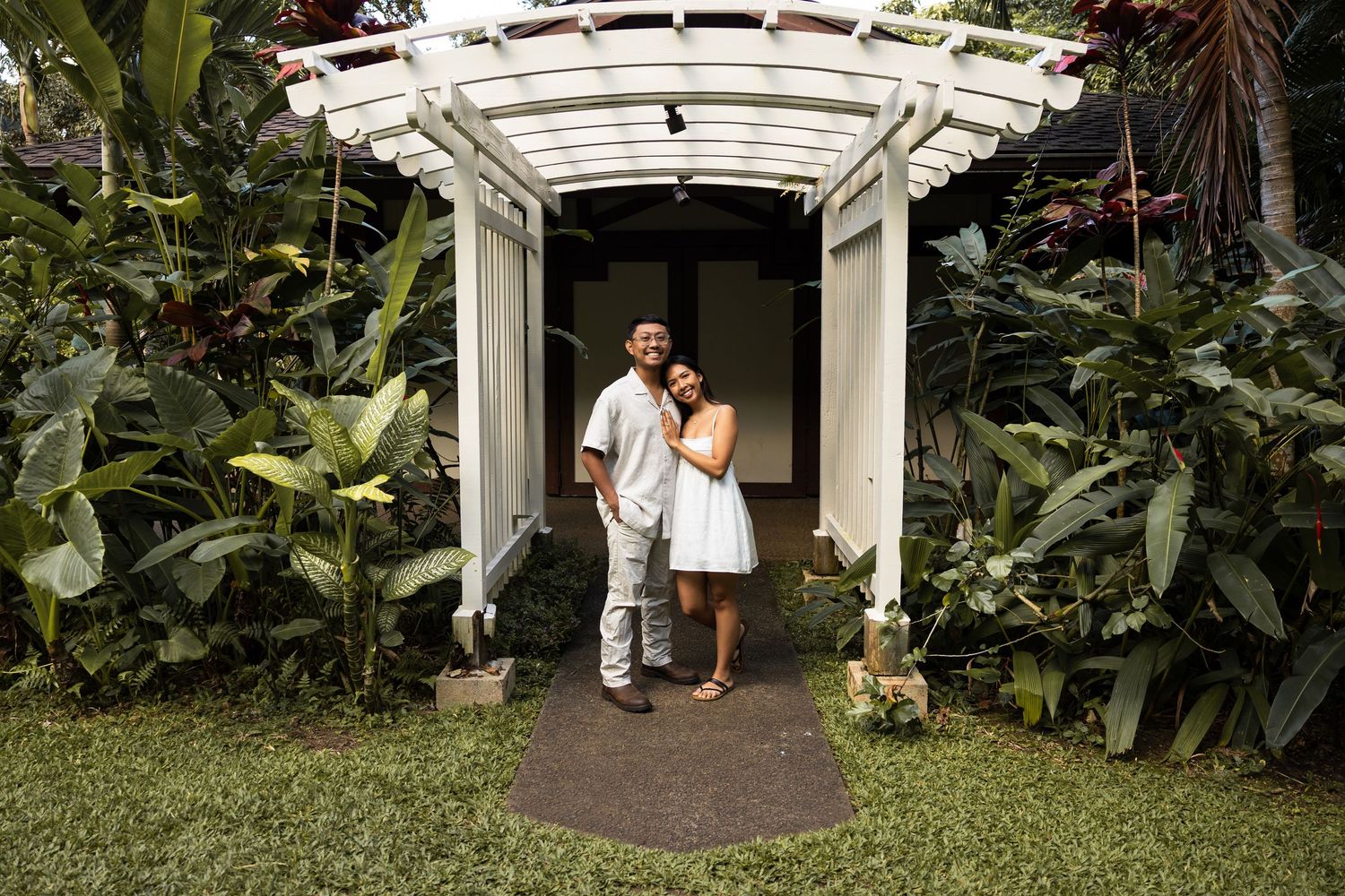 A couple stands beneath a white garden arbor surrounded by lush tropical plants.