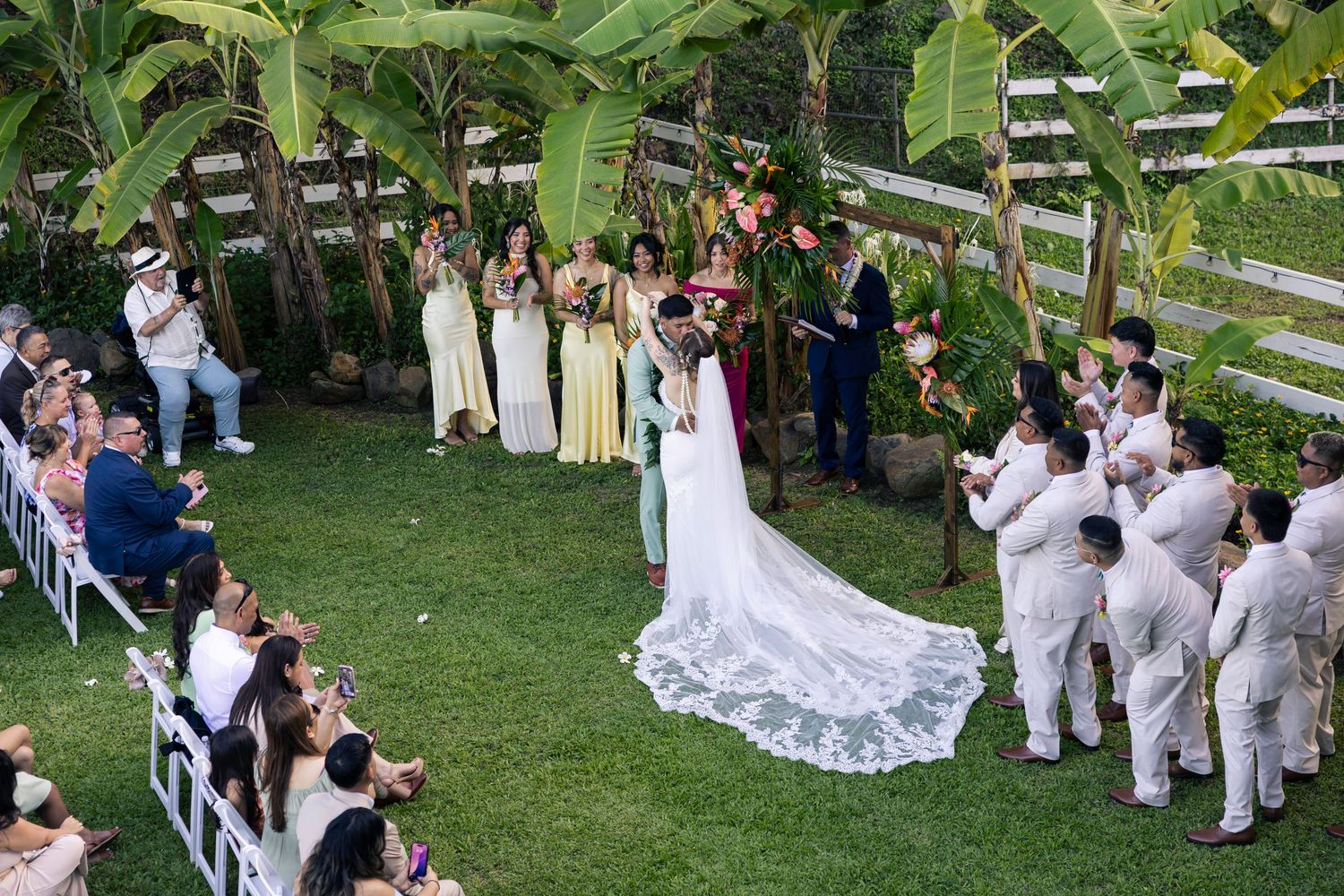 Wedding guests gather on a lush green lawn for an outdoor ceremony surrounded by tropical plants and palm trees.