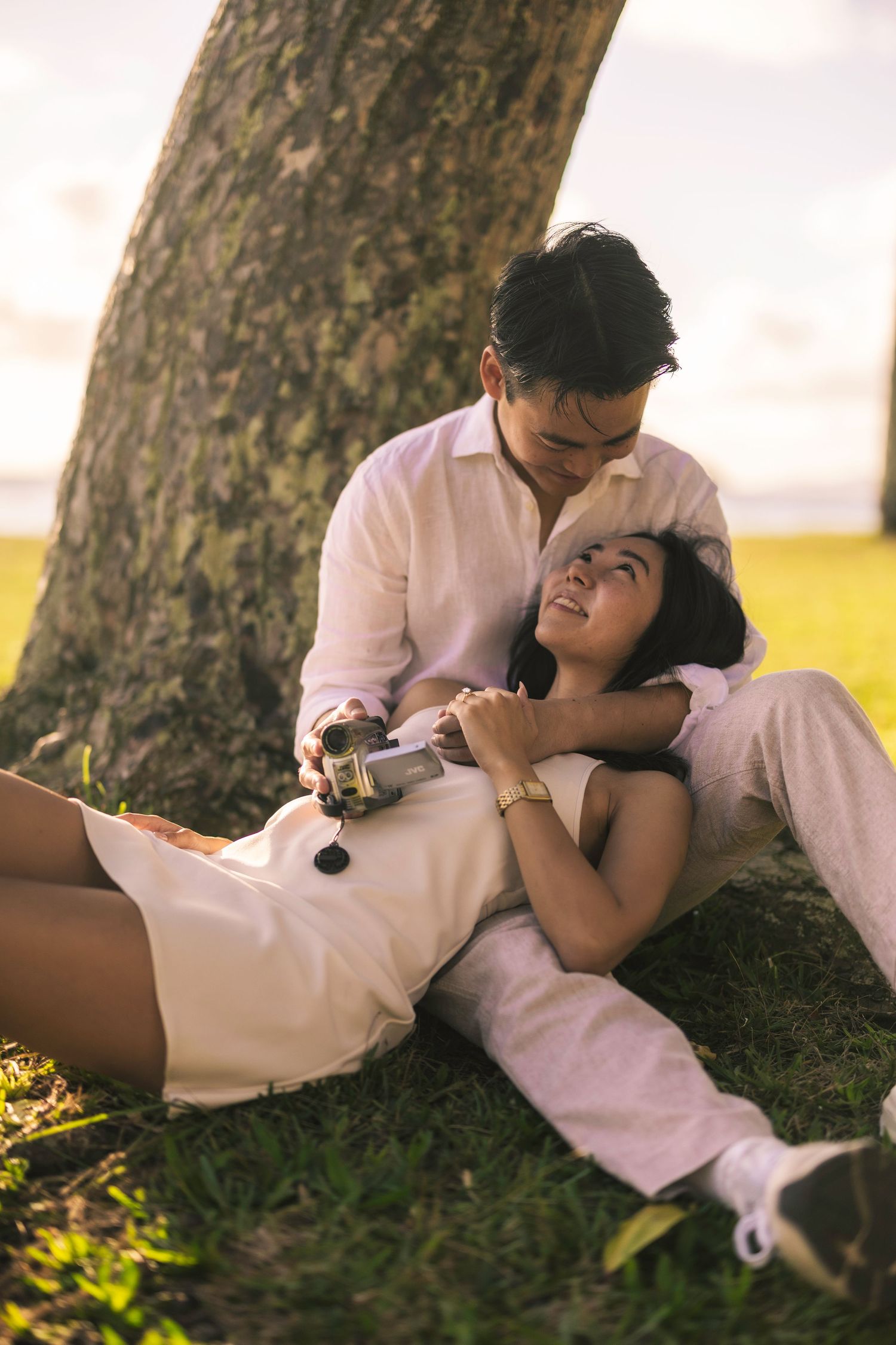 A romantic couple in white clothing embraces while lying against a tree trunk in soft sunlight.