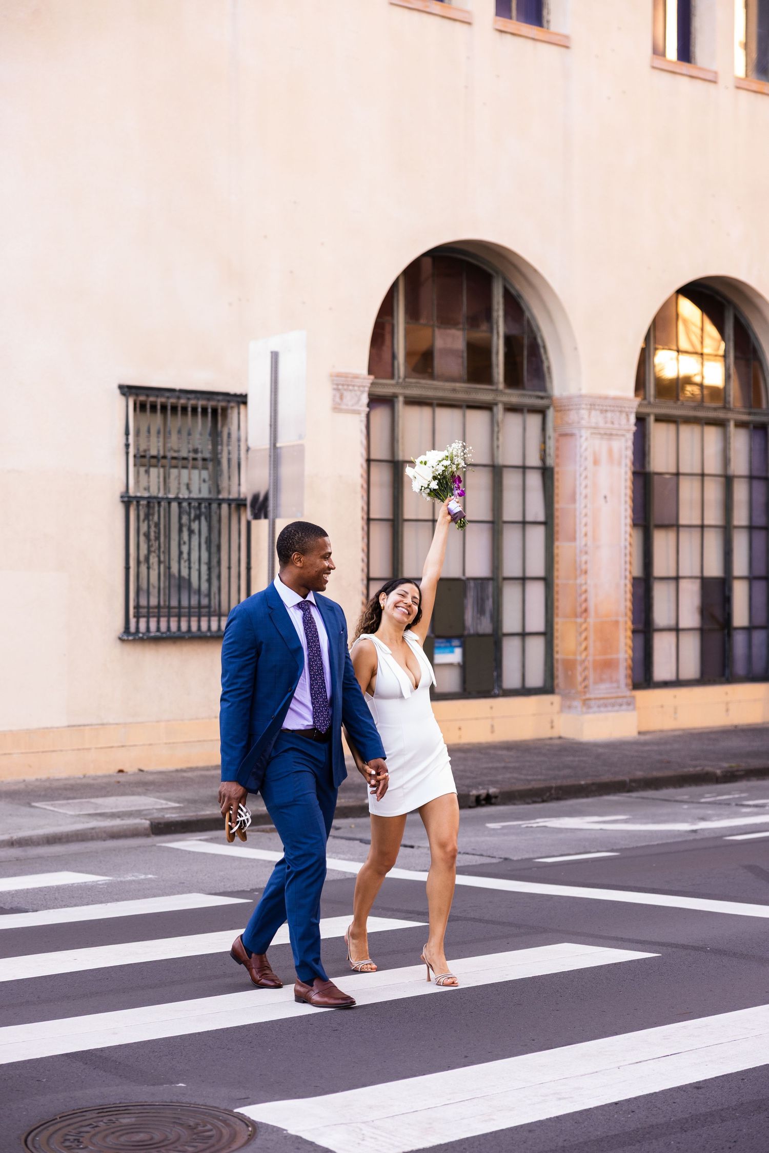 Joyful couple walking across a crosswalk, with the bride in white holding a bouquet and groom in navy suit.