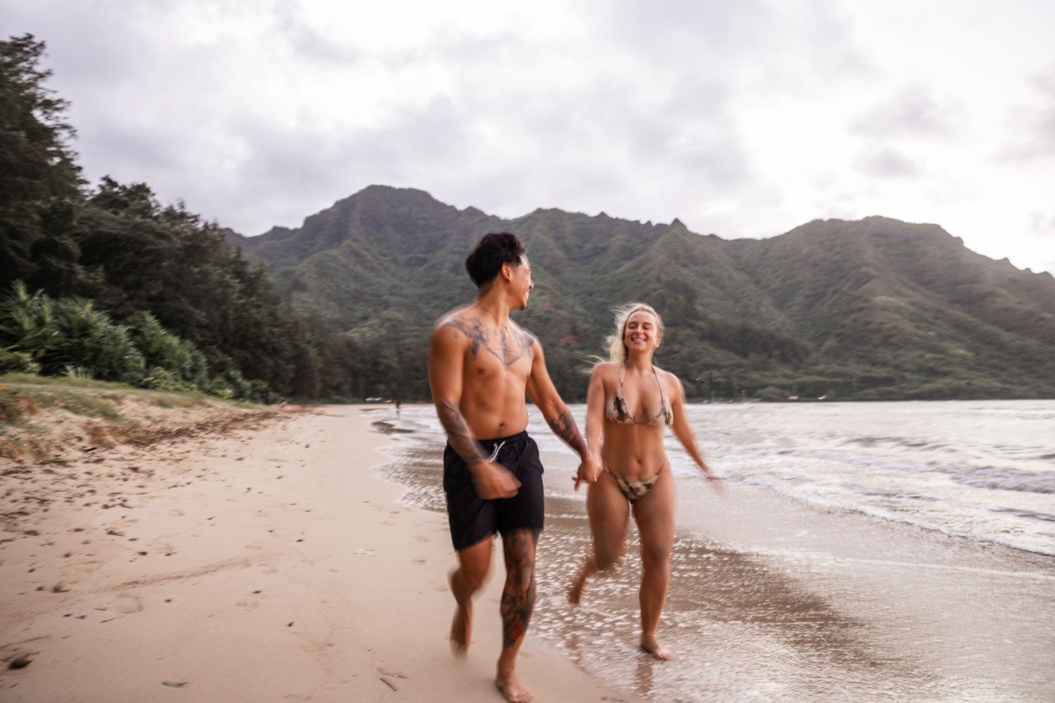 Two people walk together on a scenic beach with mountains in the background during an overcast day.