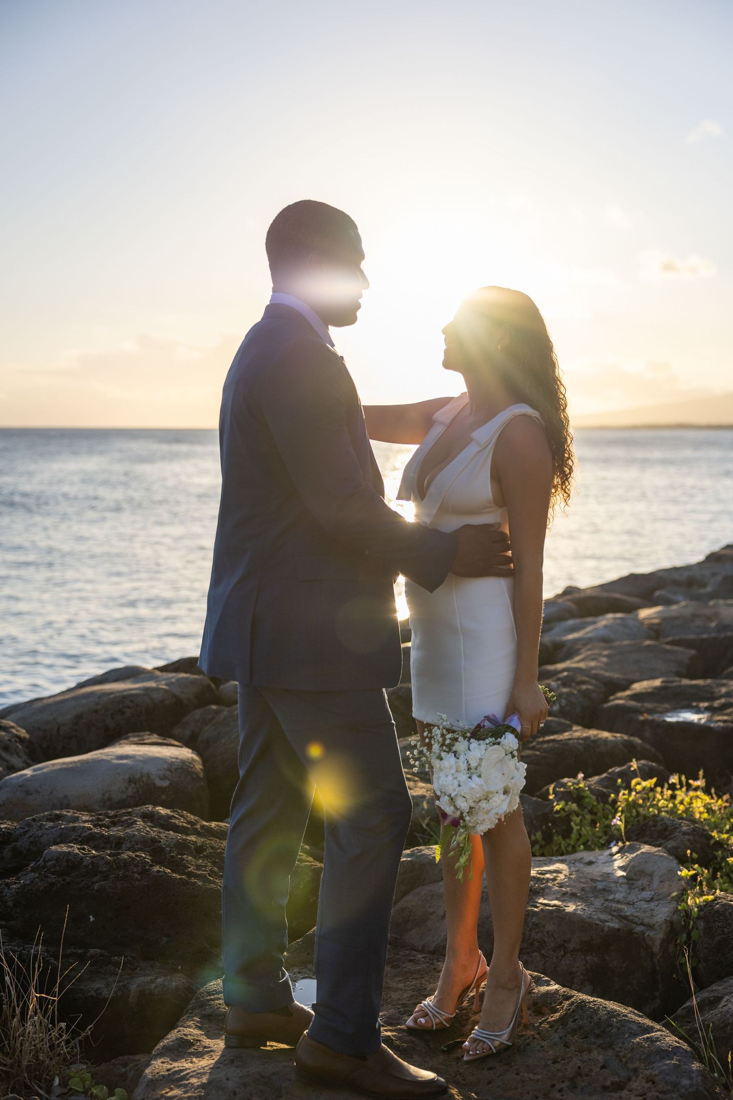Romantic sunset portrait of newlyweds embracing on rocky coastal shoreline.