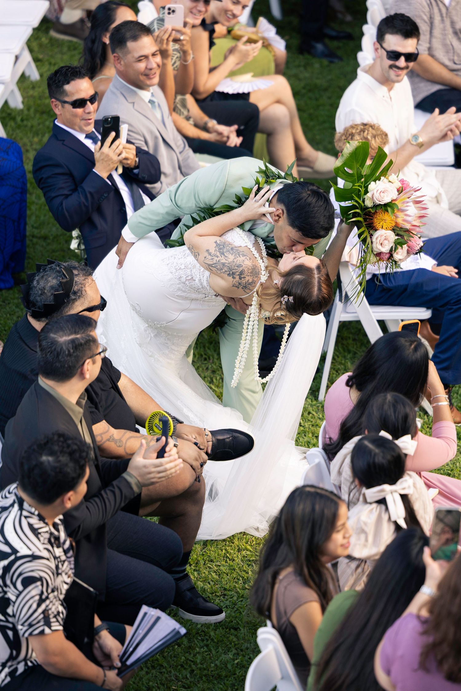 Wedding couple shares romantic kiss surrounded by guests during outdoor garden ceremony.