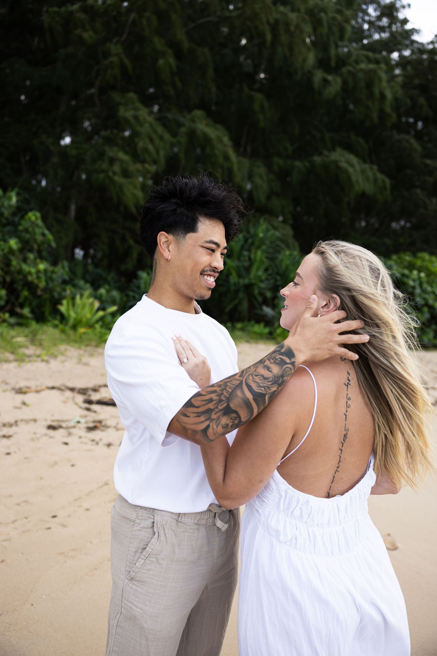 A couple laughing together on a beach with lush Hawaiian mountains creating a romantic backdrop.