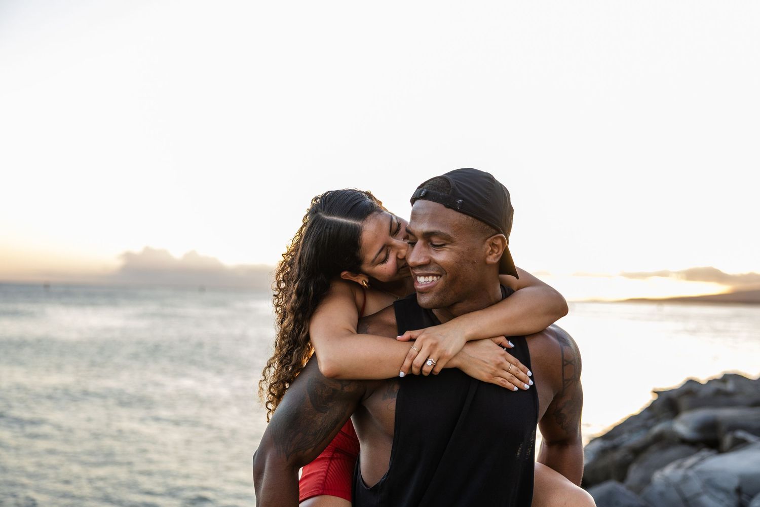 Romantic couple embraces on rocky beach at sunset with ocean waves in background.