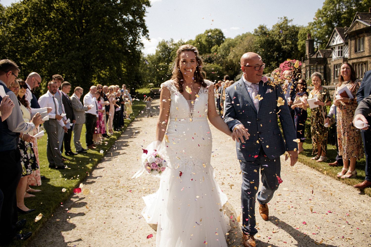 Newlyweds walk through shower of confetti on garden path lined with guests.