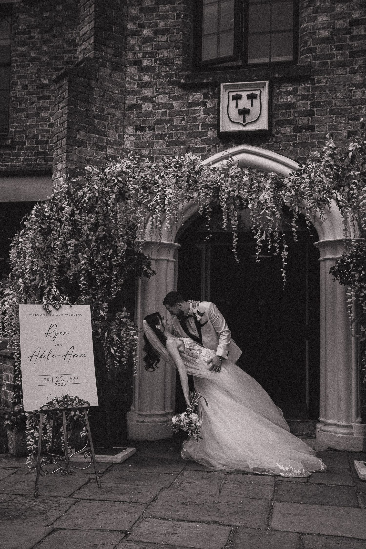Wedding couple poses under hanging floral decorations against a brick building backdrop.