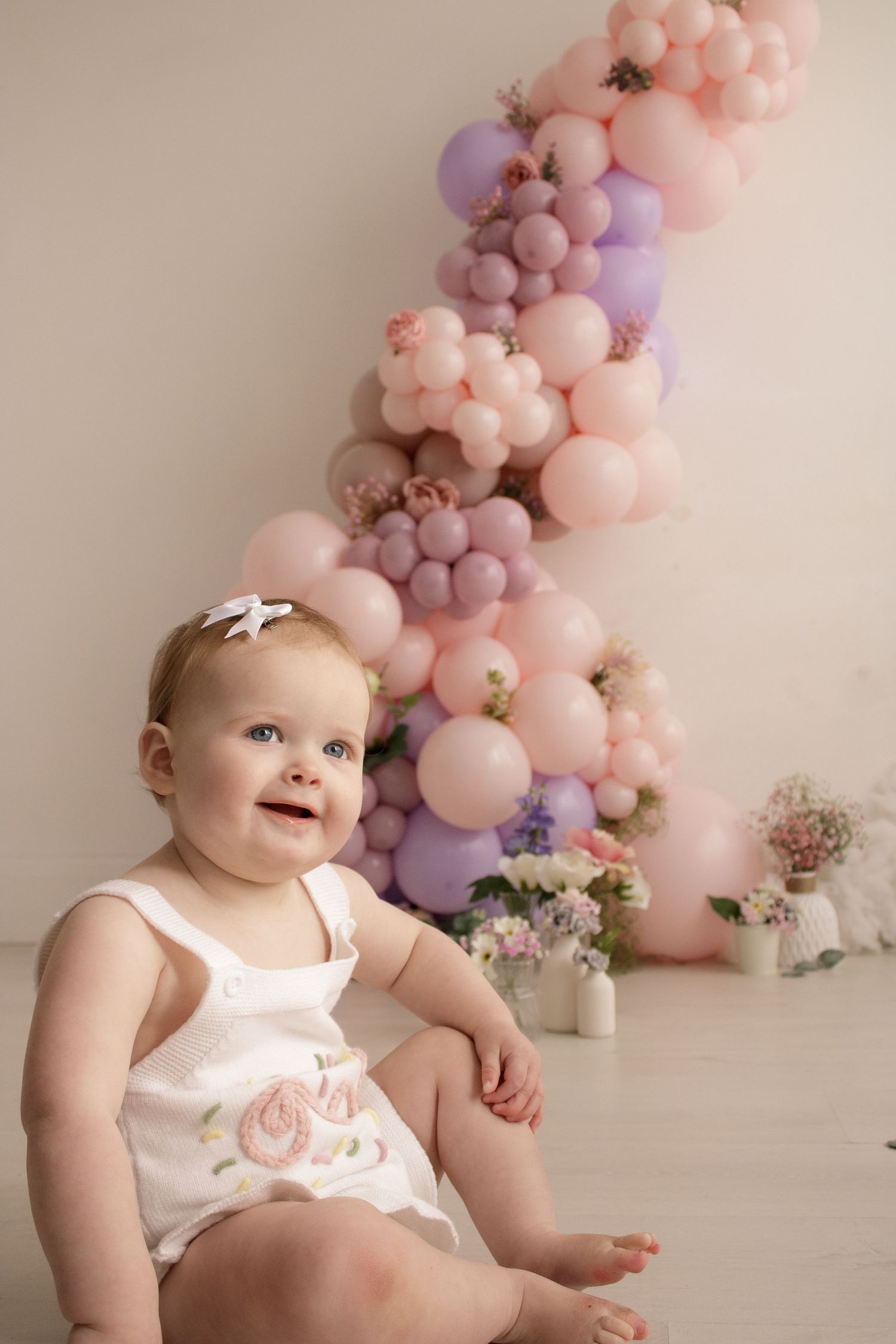 Baby in white outfit sitting against pink and purple balloon arch with floral accents.