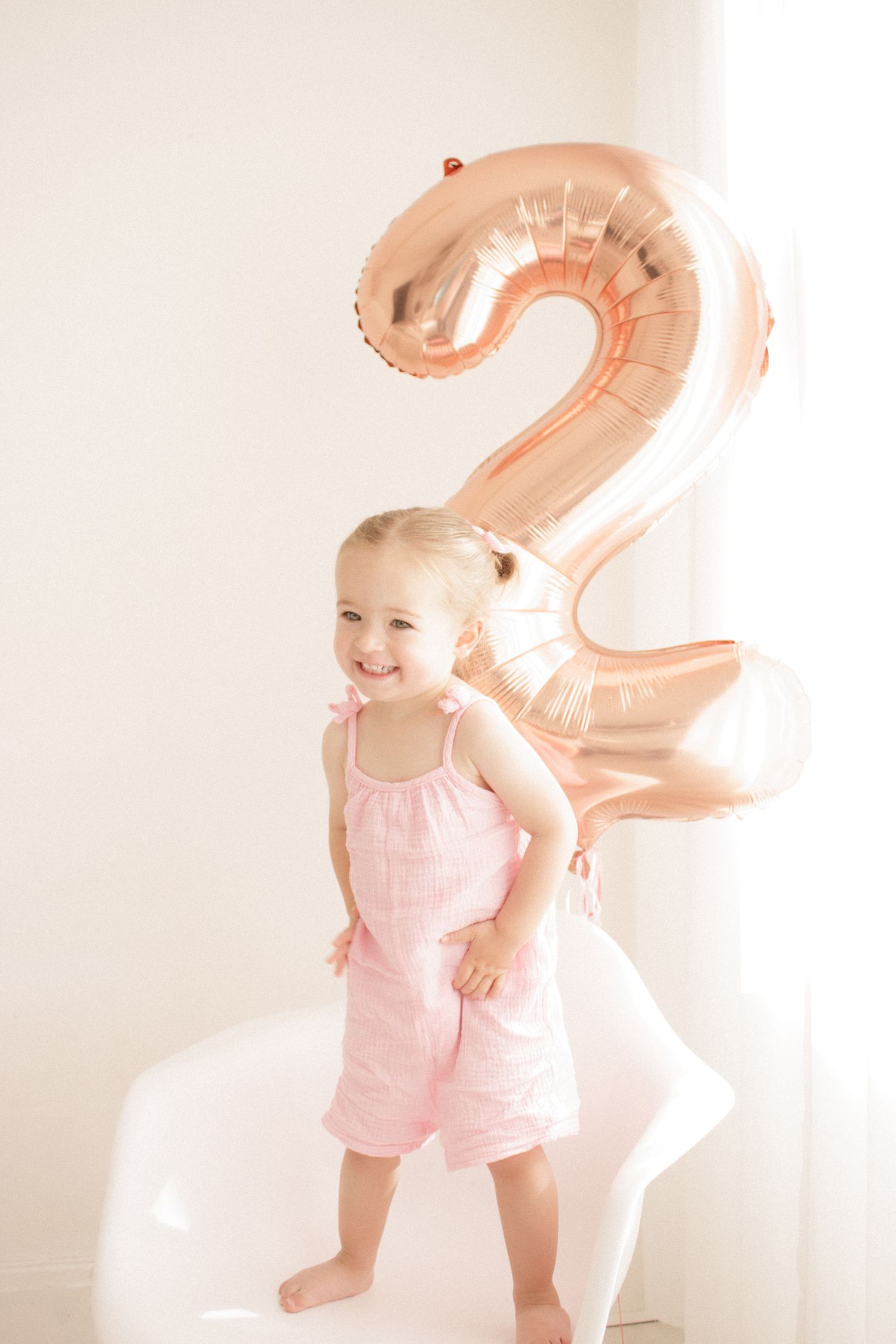 Baby in pink romper playing with rose gold number balloon in bright white room.