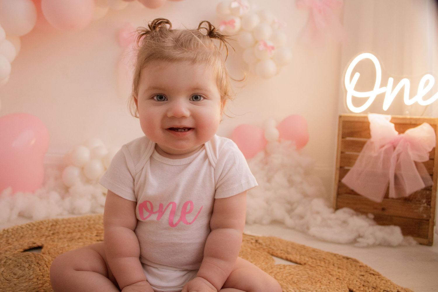 Baby in white 'One' shirt sitting against pink balloon backdrop with neon sign.
