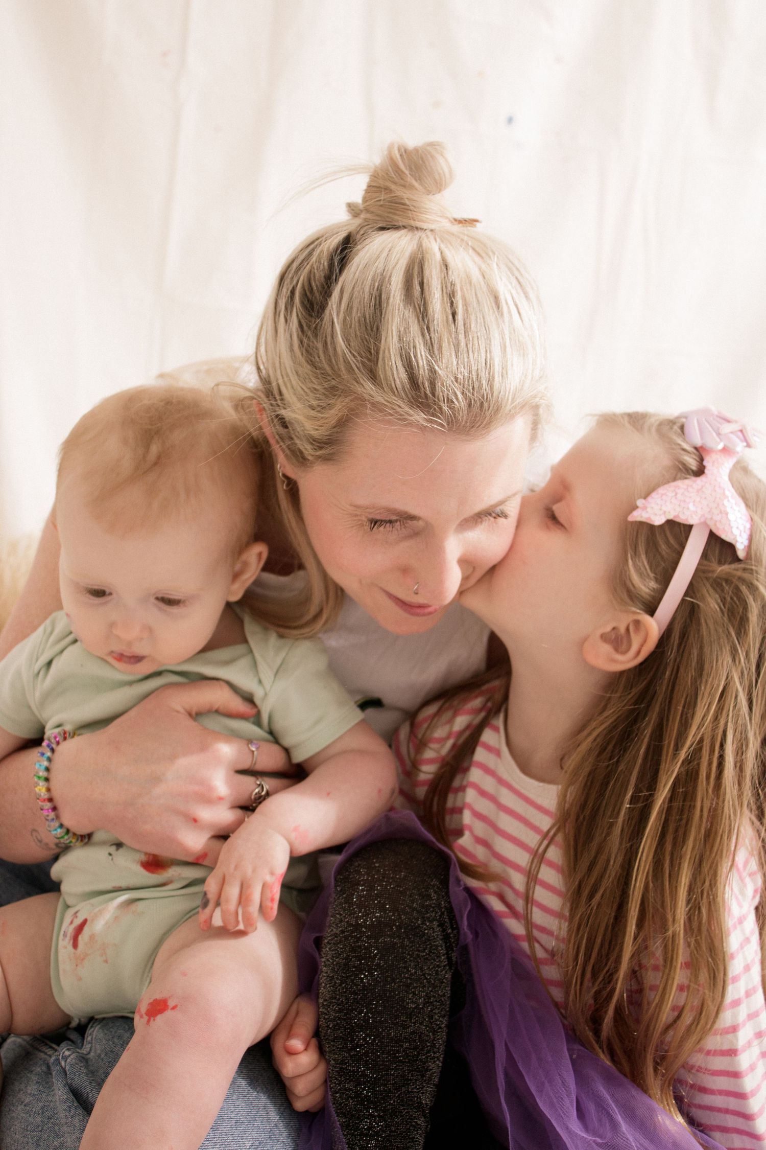 A tender family moment shows affectionate cuddles and laughter between parent and children in soft natural lighting.