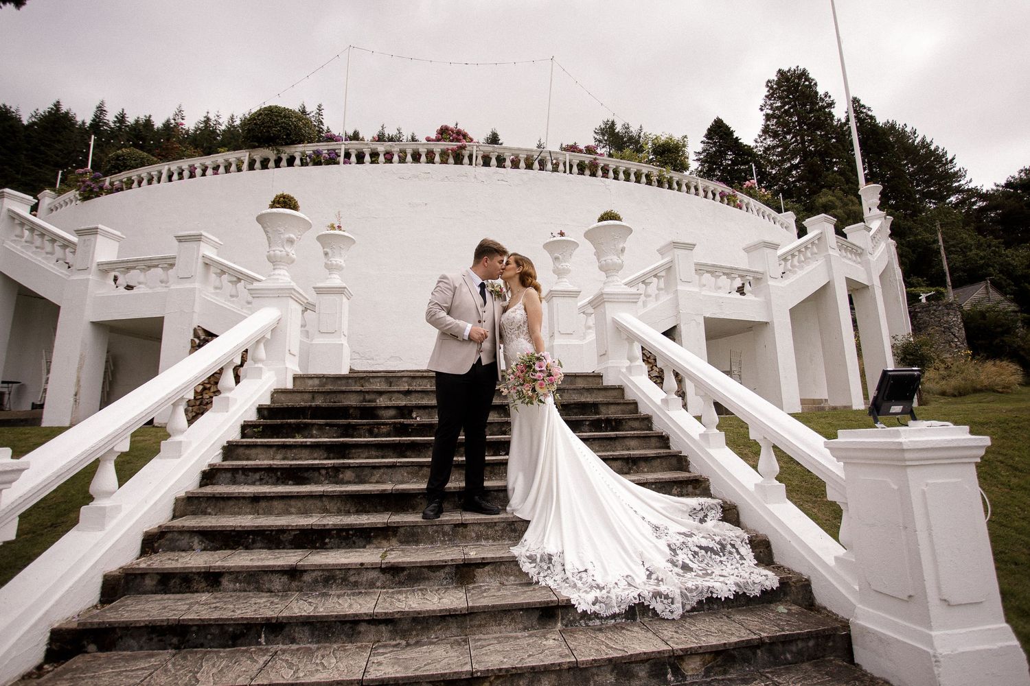 A bride in a flowing white gown twirls and poses on grand white marble stairs at an elegant wedding venue.
