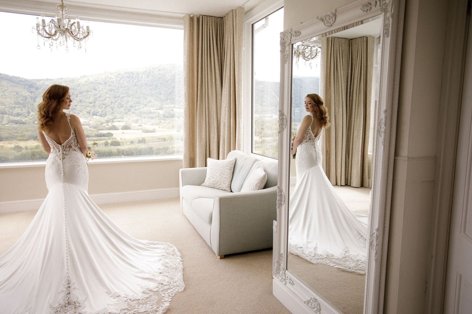 A stunning white wedding dress with intricate lace details displayed in a bright hotel room with large windows.