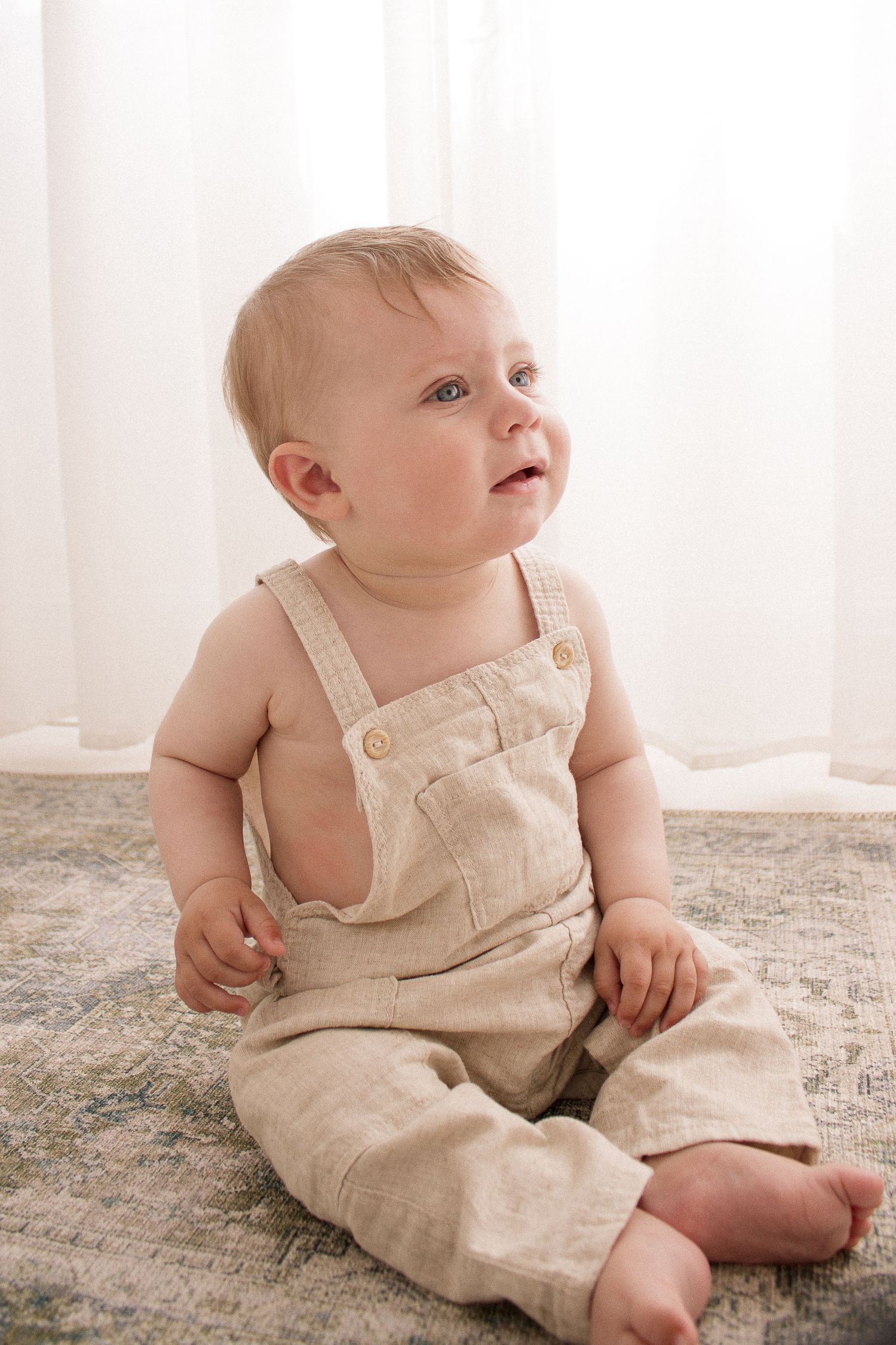 Baby wearing beige linen overalls sitting on textured carpet in natural light.