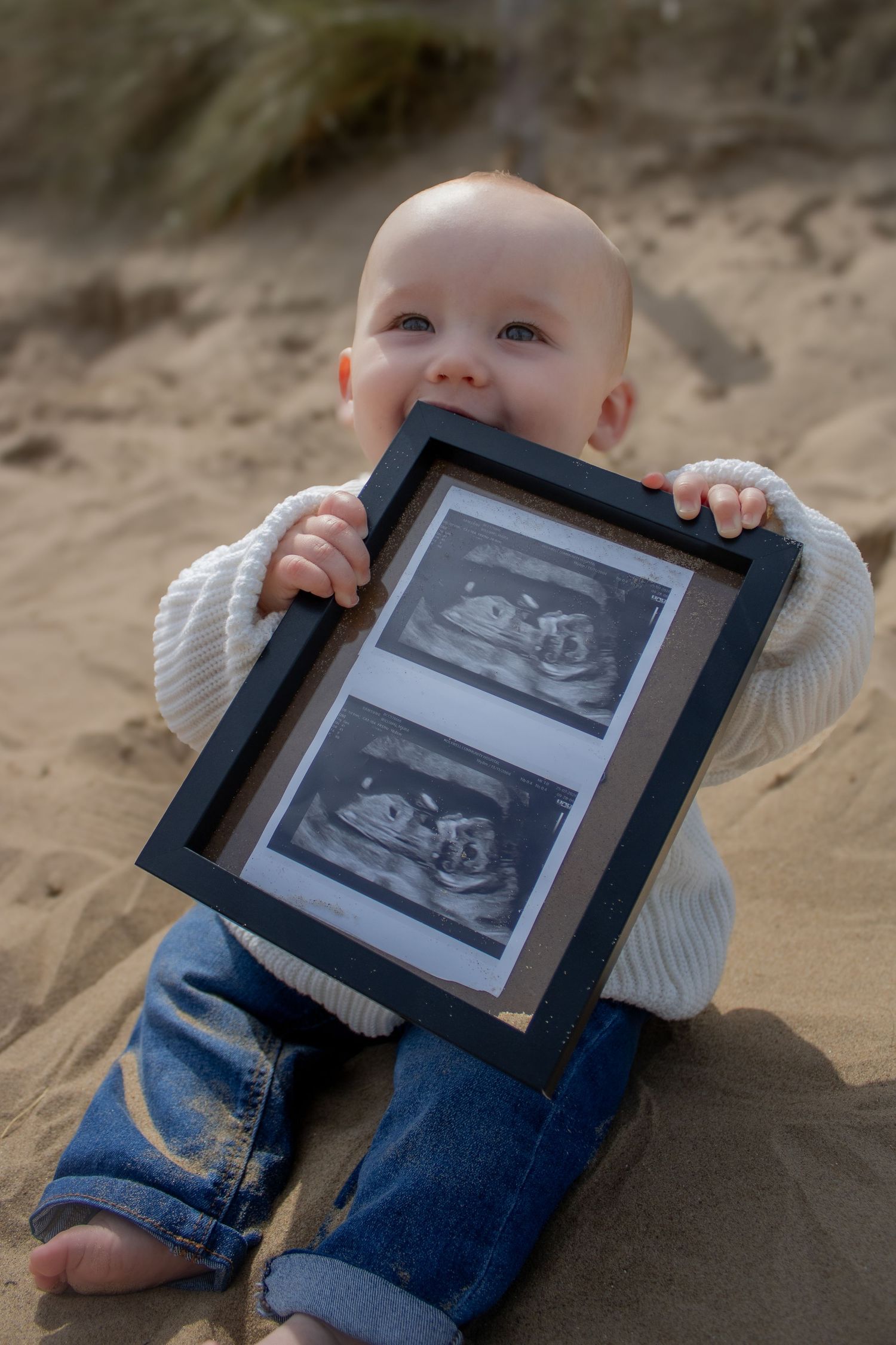 Baby in sweater and jeans holding ultrasound photos while sitting on sandy surface.
