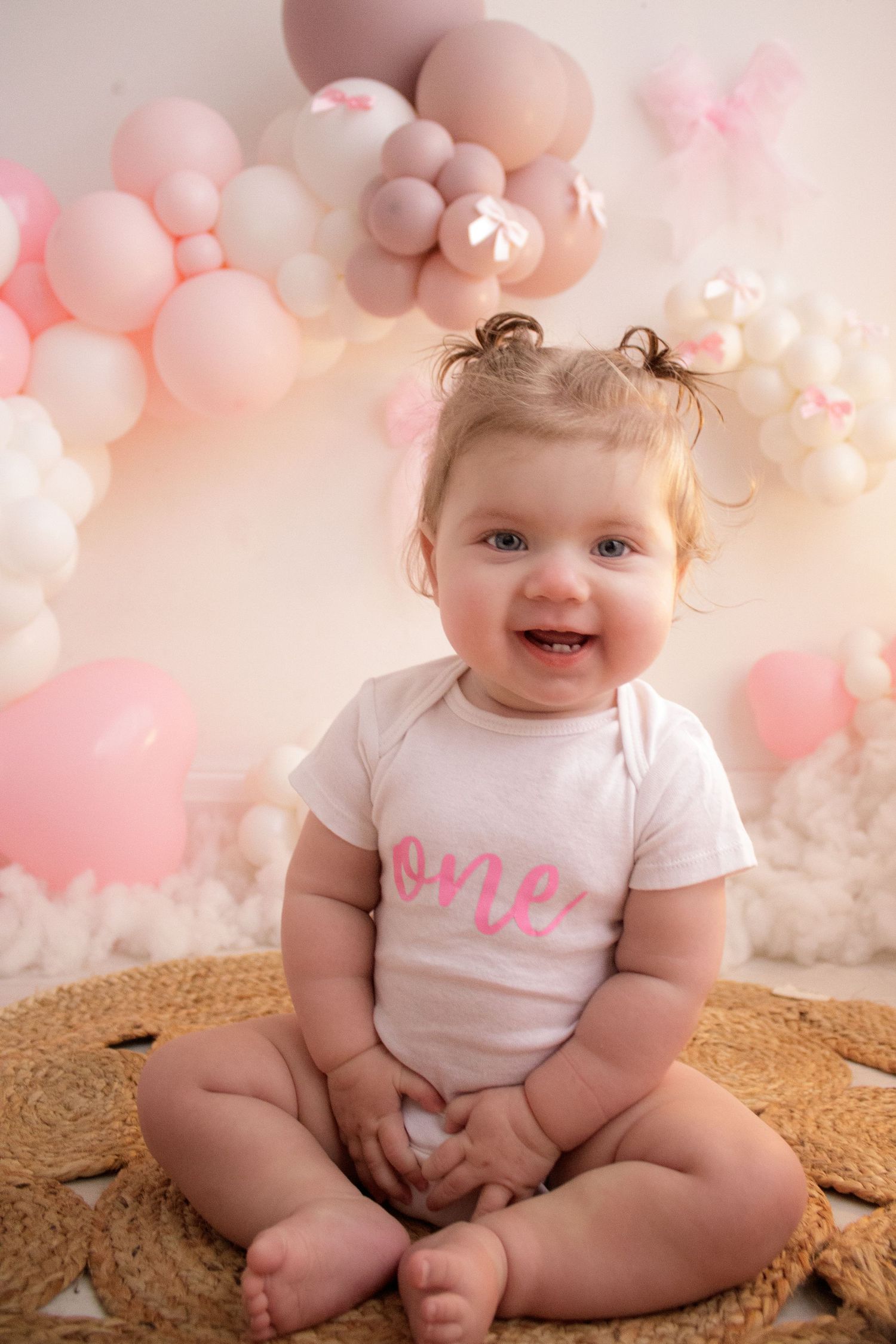 Baby sitting on round rug with pink balloon arch and fairy lights in background.