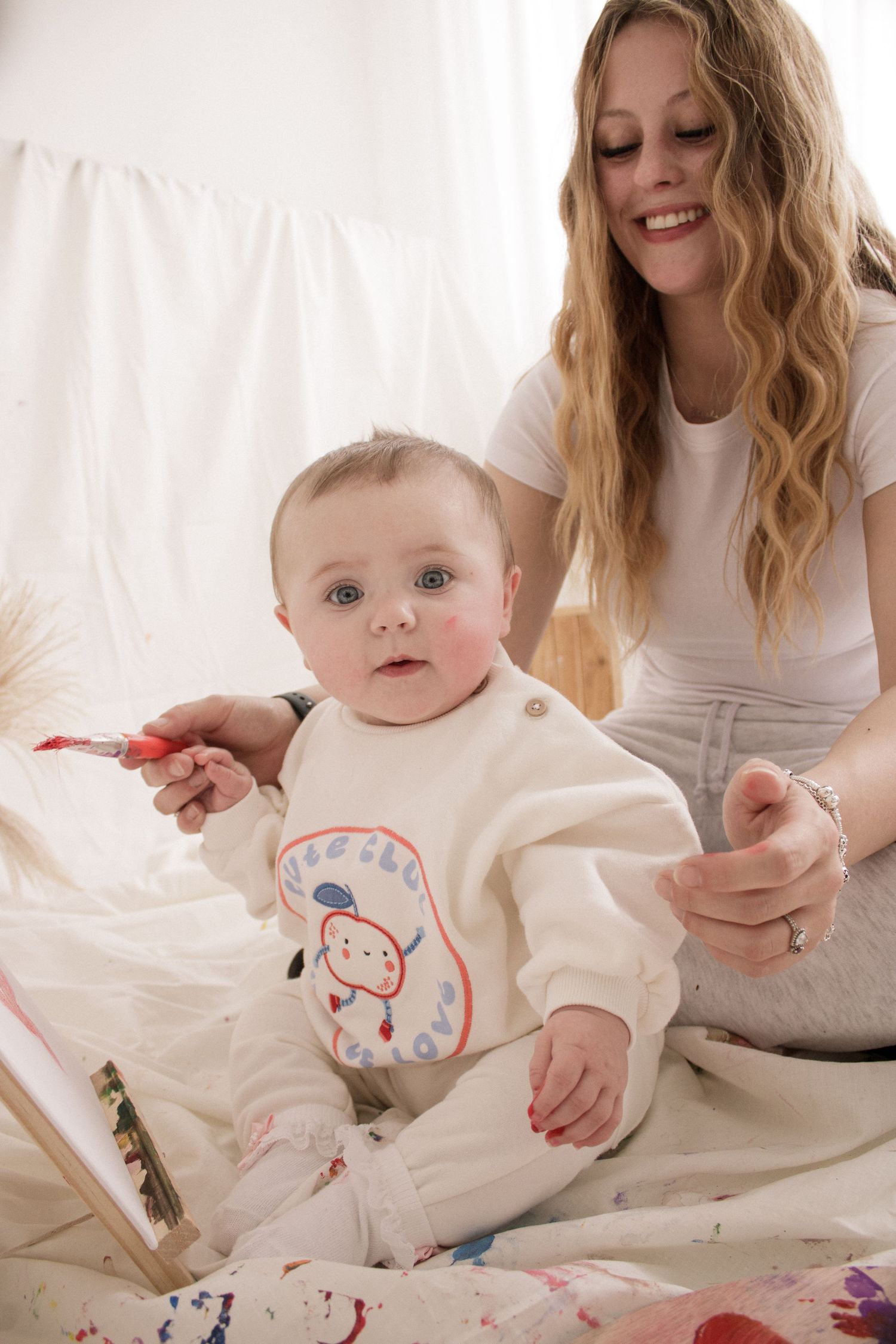 A baby in white clothing sits on a bed with colorful drawings while being assisted by someone in white.