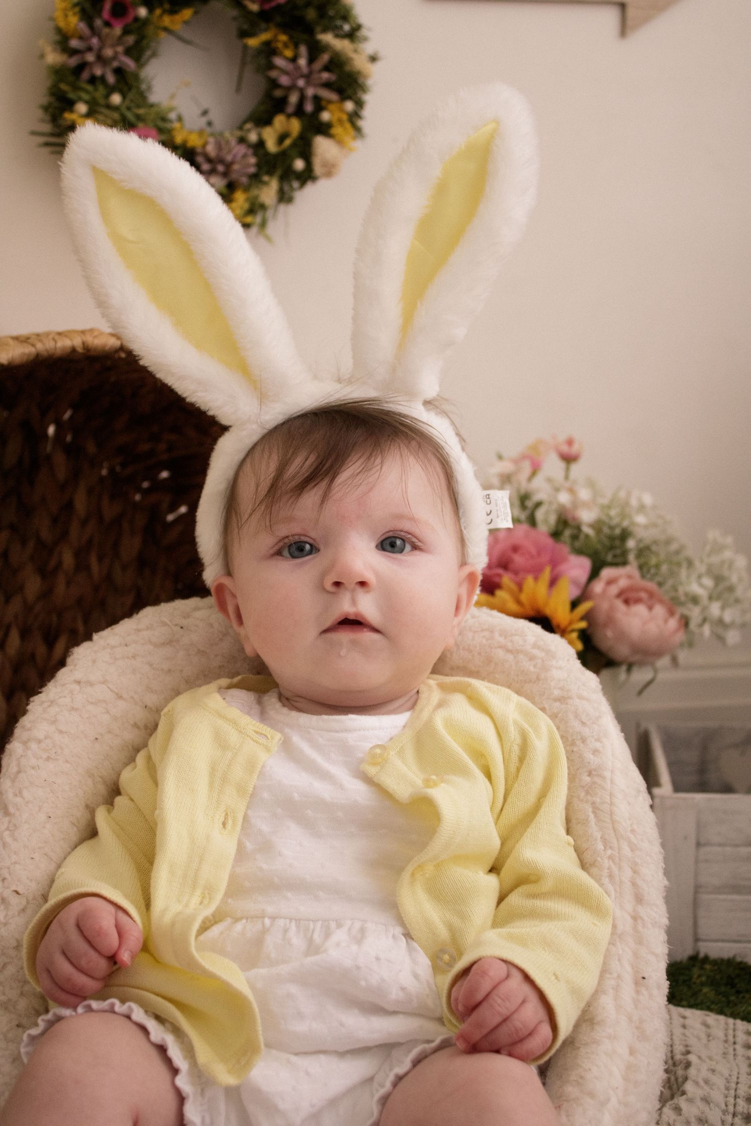 Baby wearing bunny ears and yellow cardigan sitting in plush chair with spring decorations.