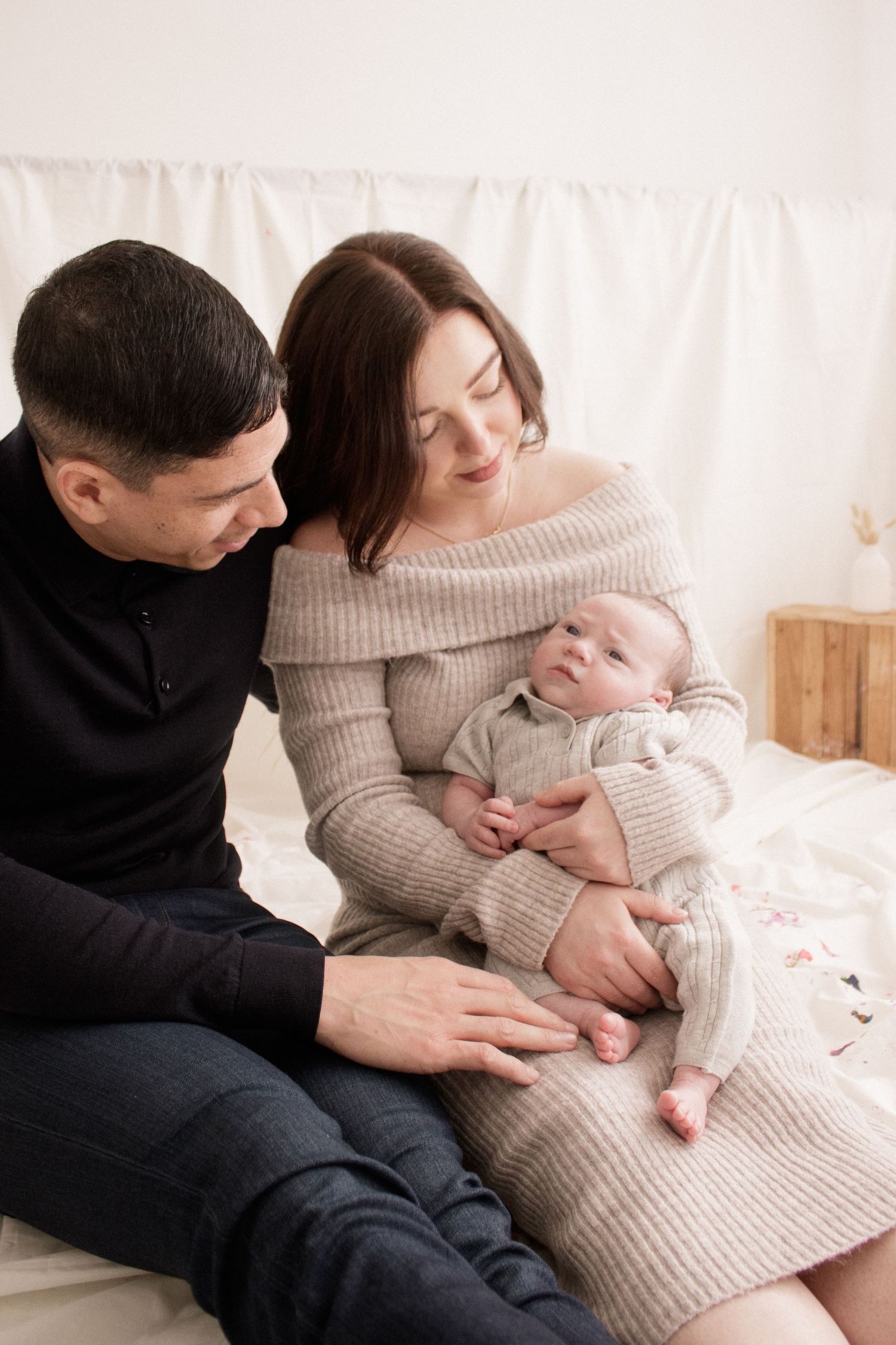 A couple in casual attire sits together on a light-colored couch while holding and gazing at their newborn baby.