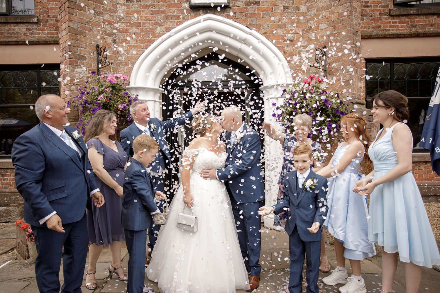 Wedding party celebrating with confetti and bubbles outside a rustic brick building.