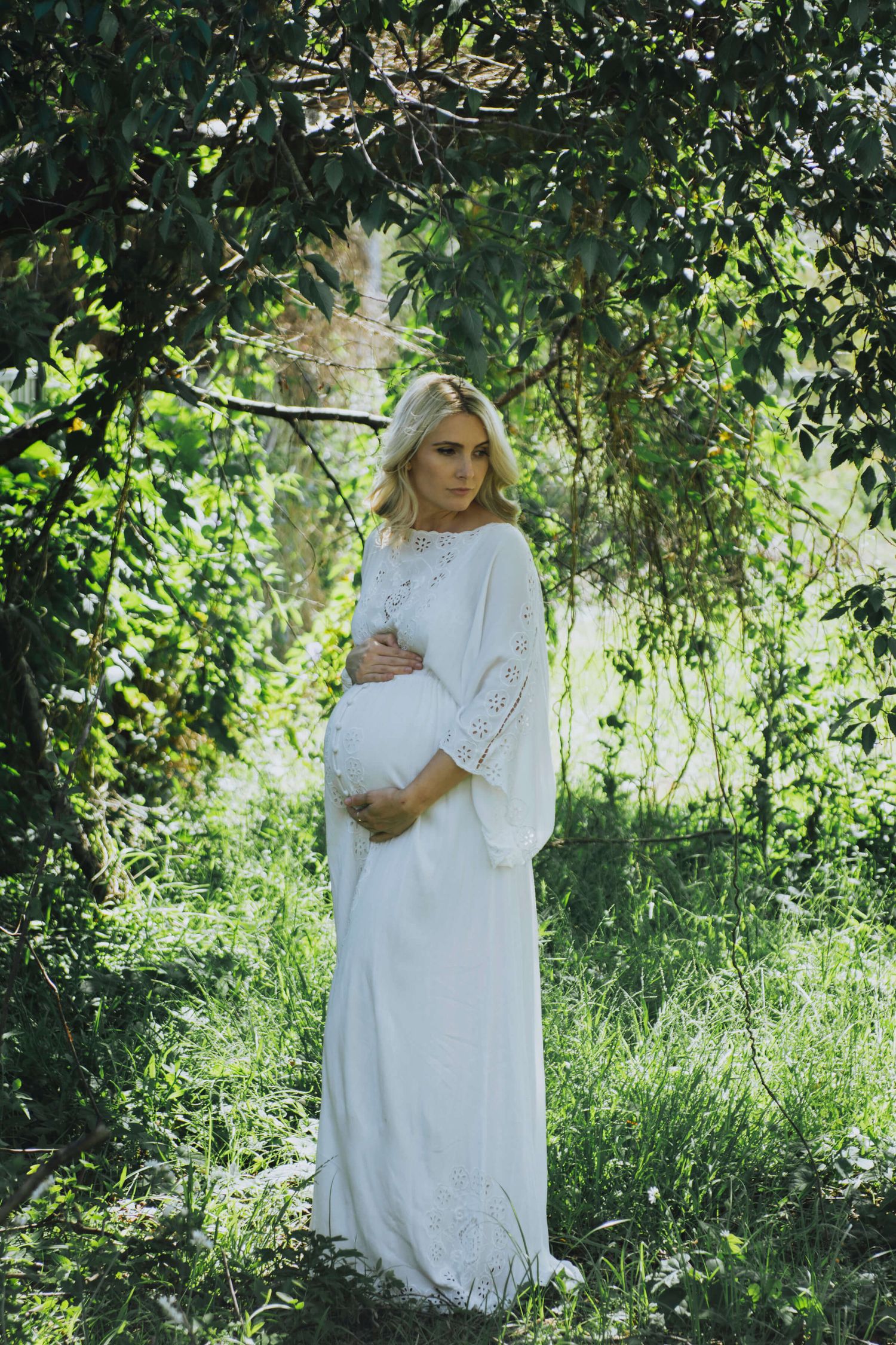 Pregnant woman holding belly surrounded by trees and overgrown bush during at-home session - Zara King Photography, Sydney Australia