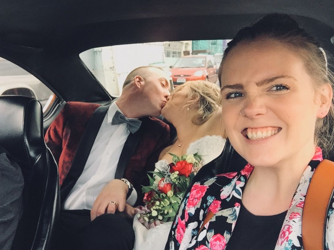 Zara King smiling in the front seat while a newly married couple kiss in the back seat - Zara King Photography, Dunedin NZ
