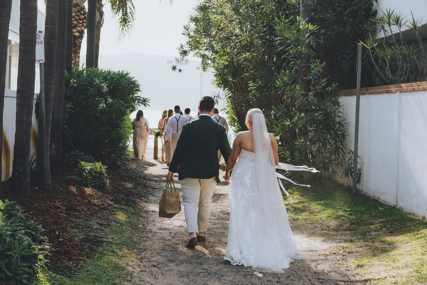 newlywed couple holding hands and a picnic basket on their way to a beach with their bridal party - Zara King Photography, Sydney Australia
