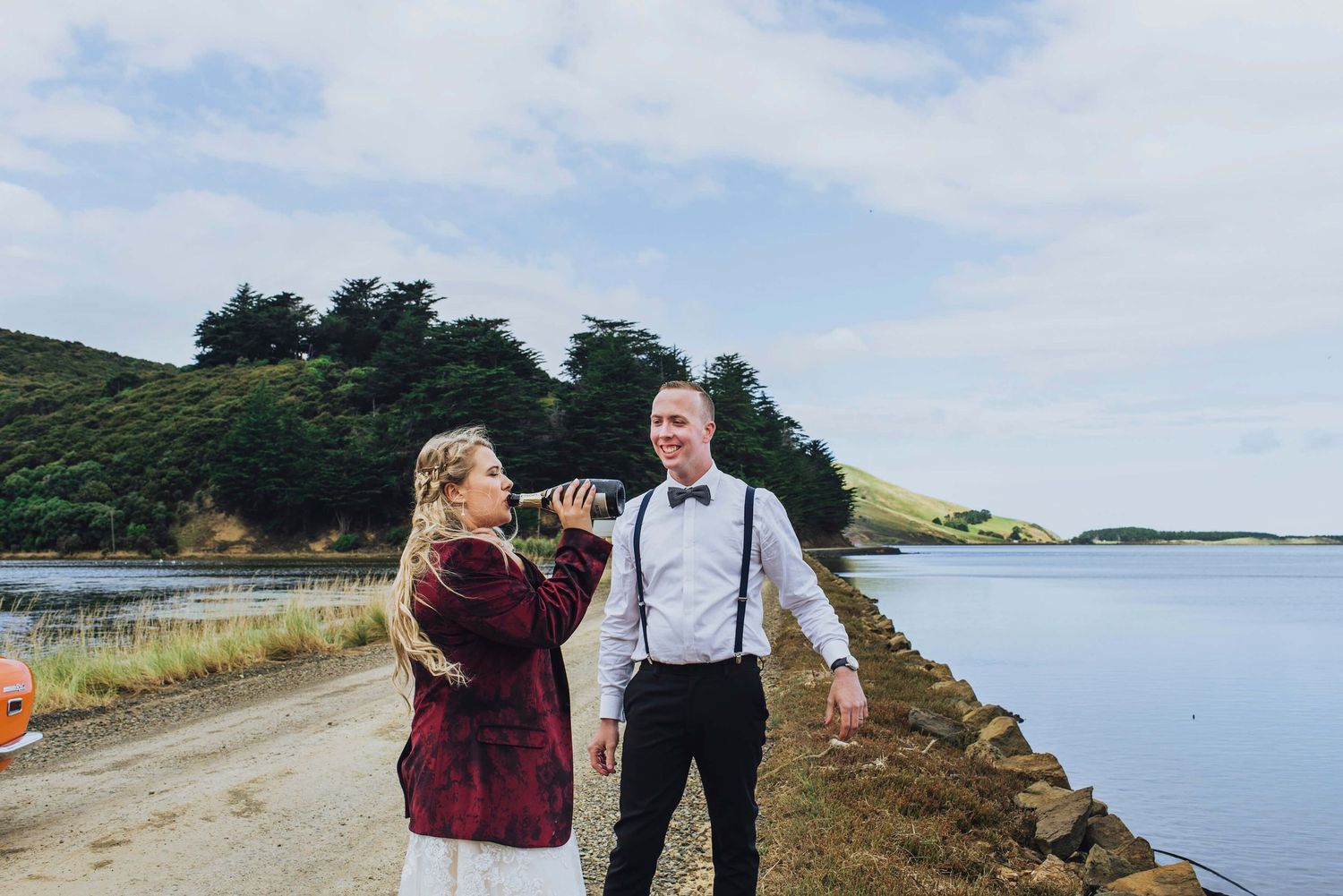 Bride drinking straight from the wine bottle while groom smiles at her in Dunedin, NZ - Zara King Photography