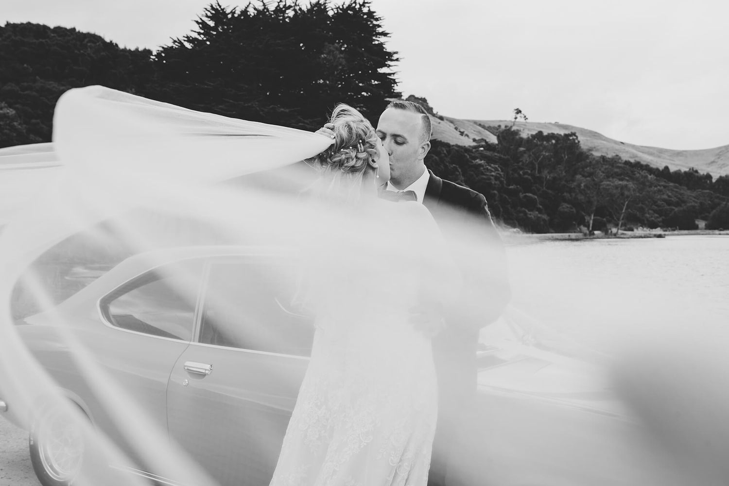Bride and groom kissing while sitting on car with bride's veil blowing in the wind in Dunedin, NZ - Zara King Photography