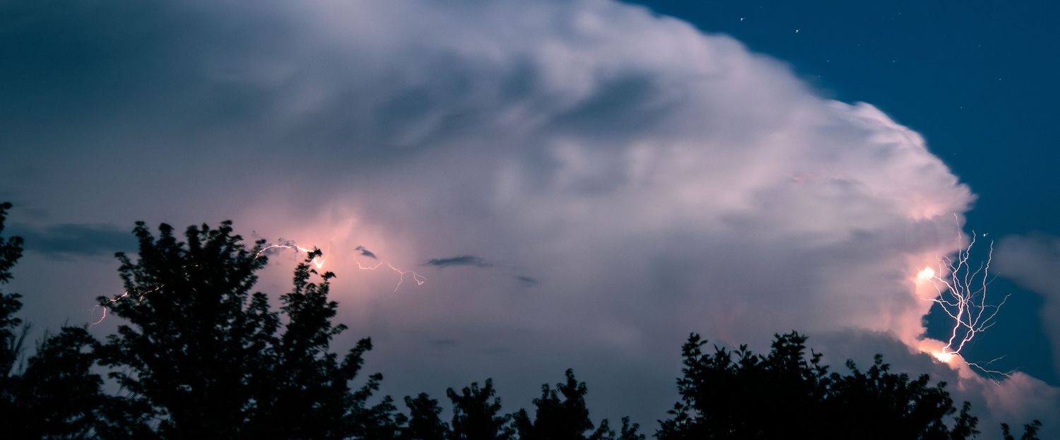 Wide panoramic view of lightning flashing inside a large storm cloud above dark tree silhouettes.