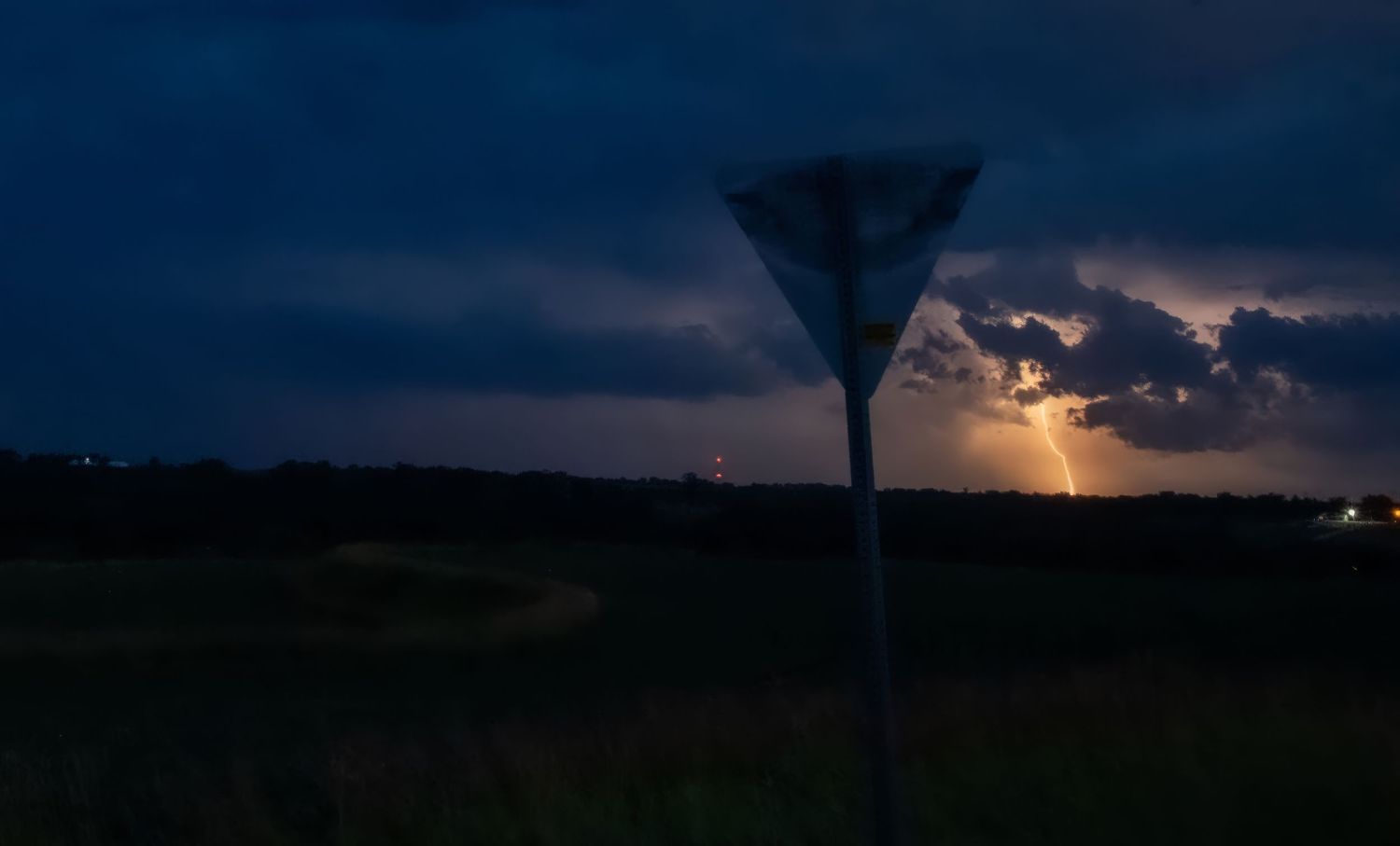 Lightning illuminates storm clouds behind a roadside sign in a dark open field at night.