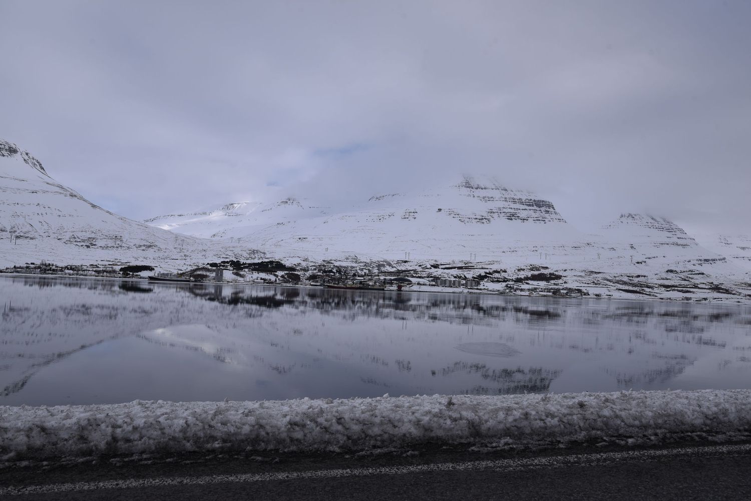 Snow-covered mountains reflect perfectly in a calm fjord under overcast skies in Iceland.
