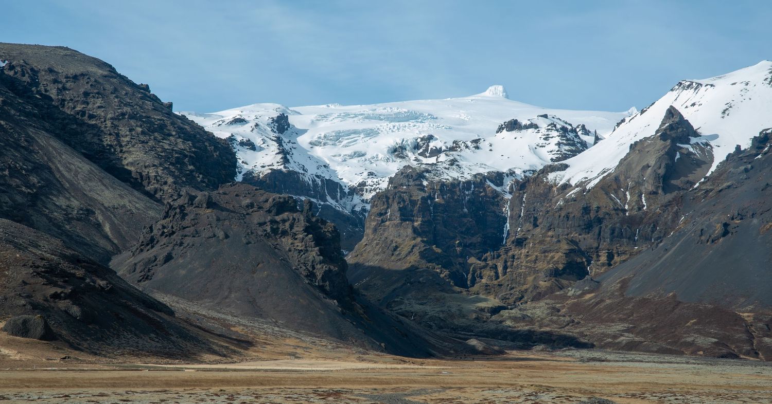 Snow-capped Icelandic mountains tower over a rugged volcanic landscape under a clear blue sky.