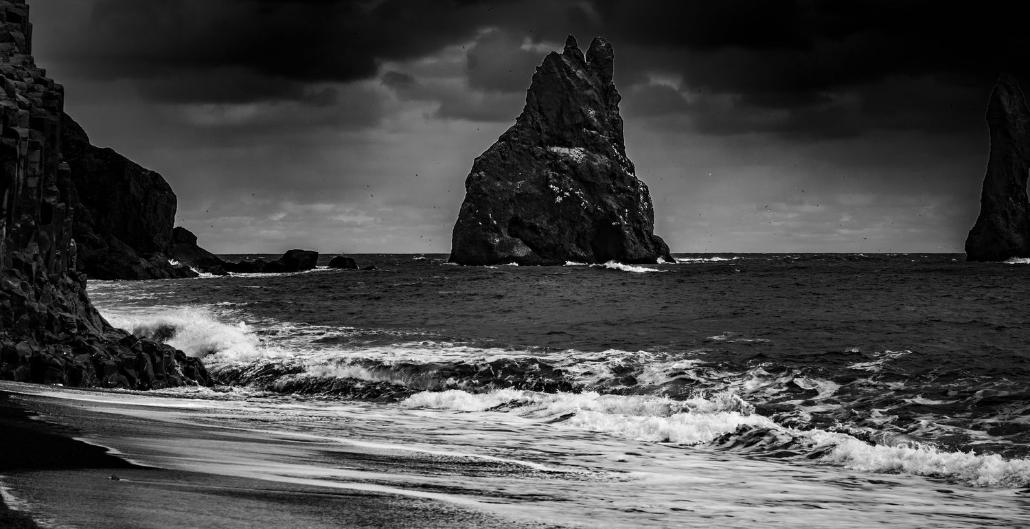 Black and white photo of a jagged sea stack rising from turbulent ocean waves on a dark sandy beach.