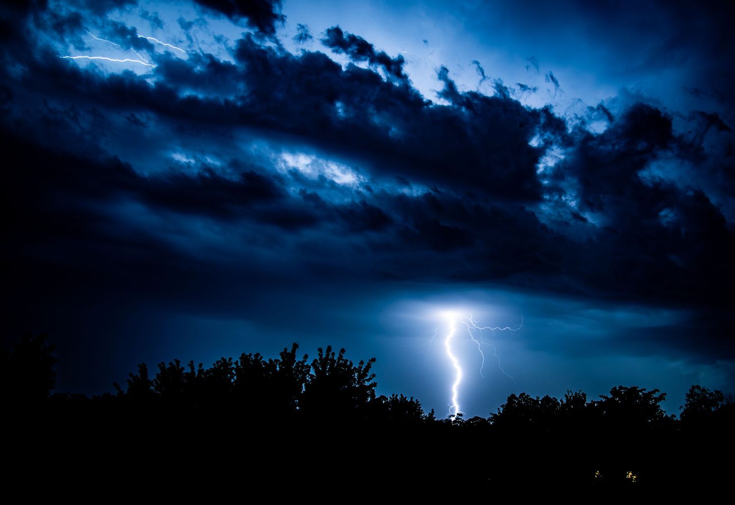 Brilliant lightning bolt illuminating deep blue storm clouds over silhouetted trees at night.