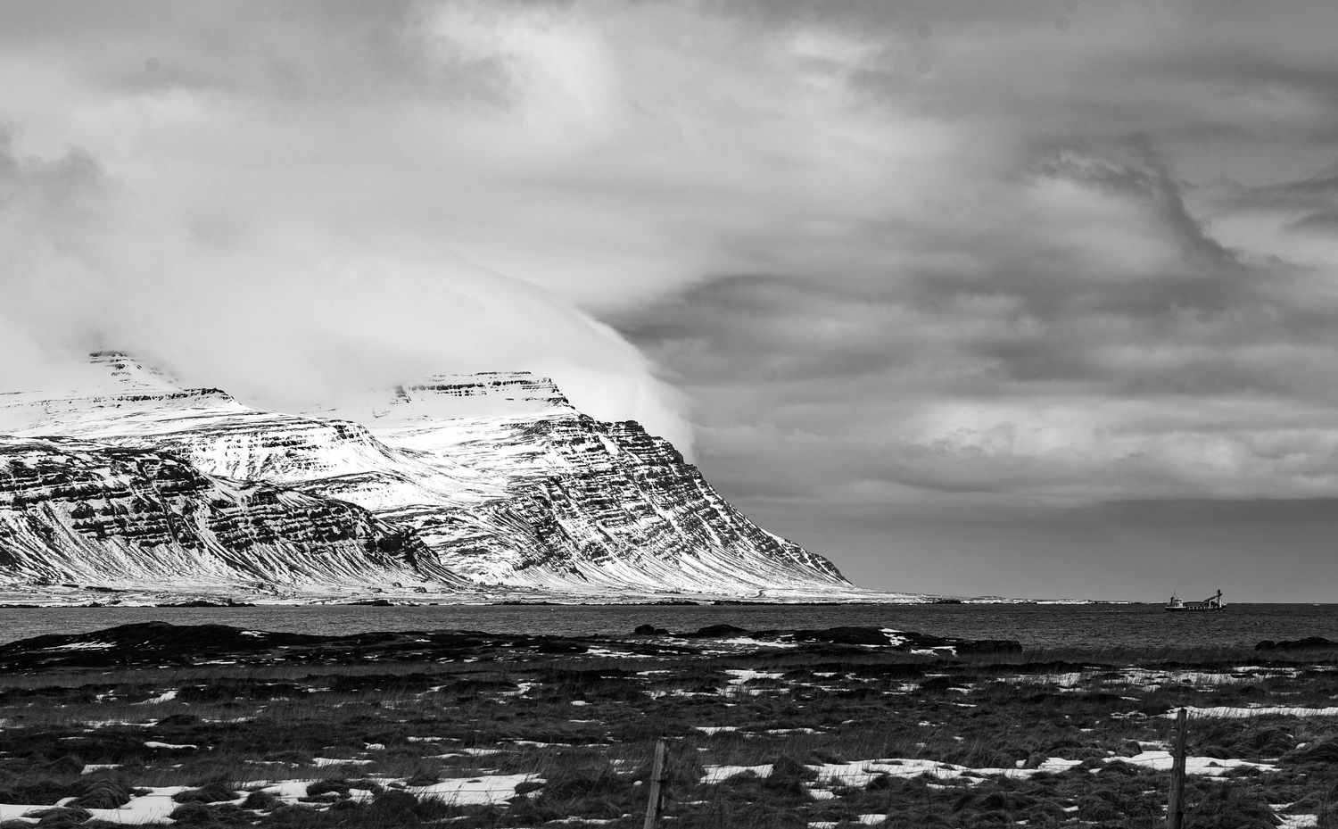 Black and white panoramic view of snow-covered Icelandic mountain range reflecting on rocky coastal shoreline.