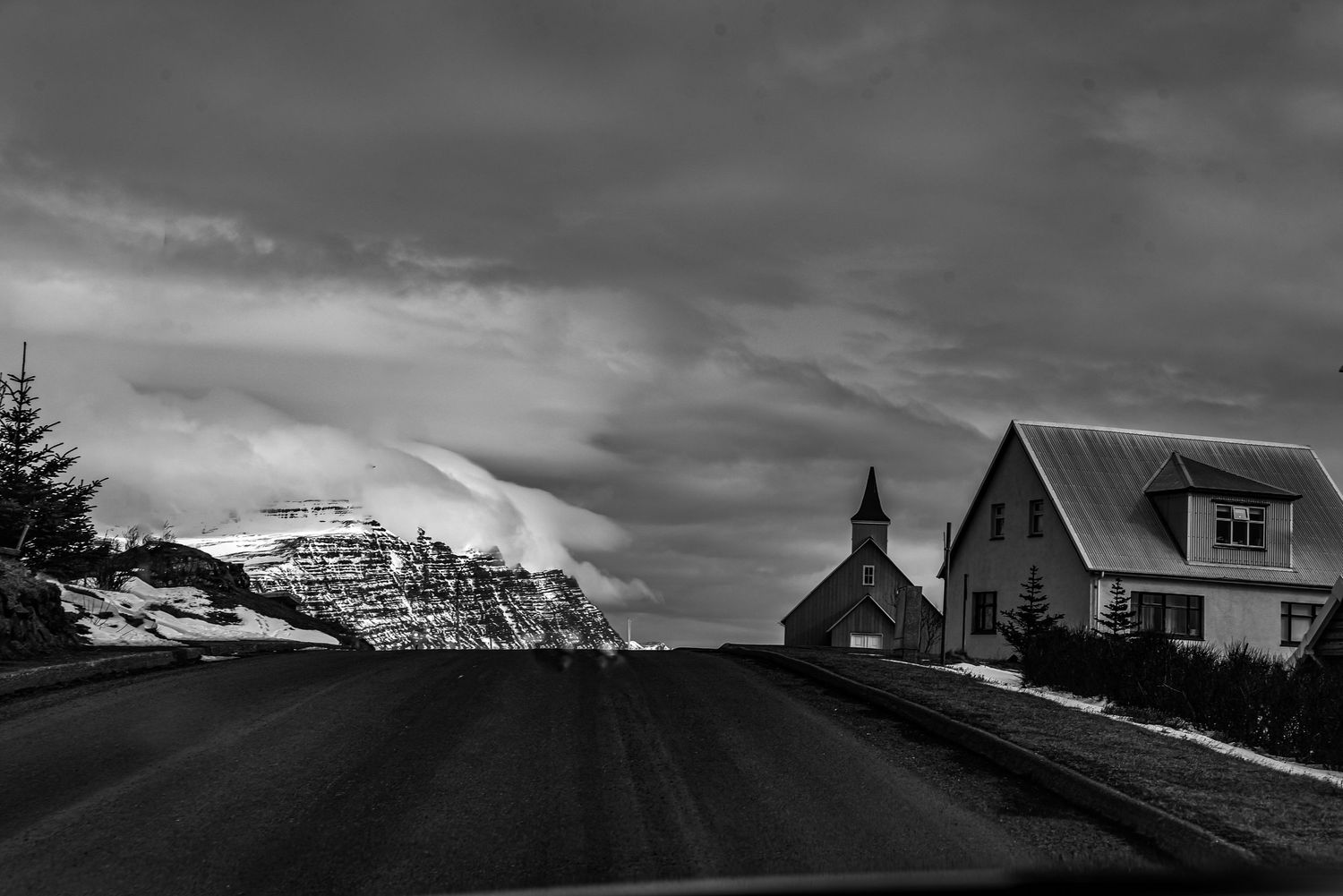 Black and white road leading to a church and house with dramatic snowy mountains behind.