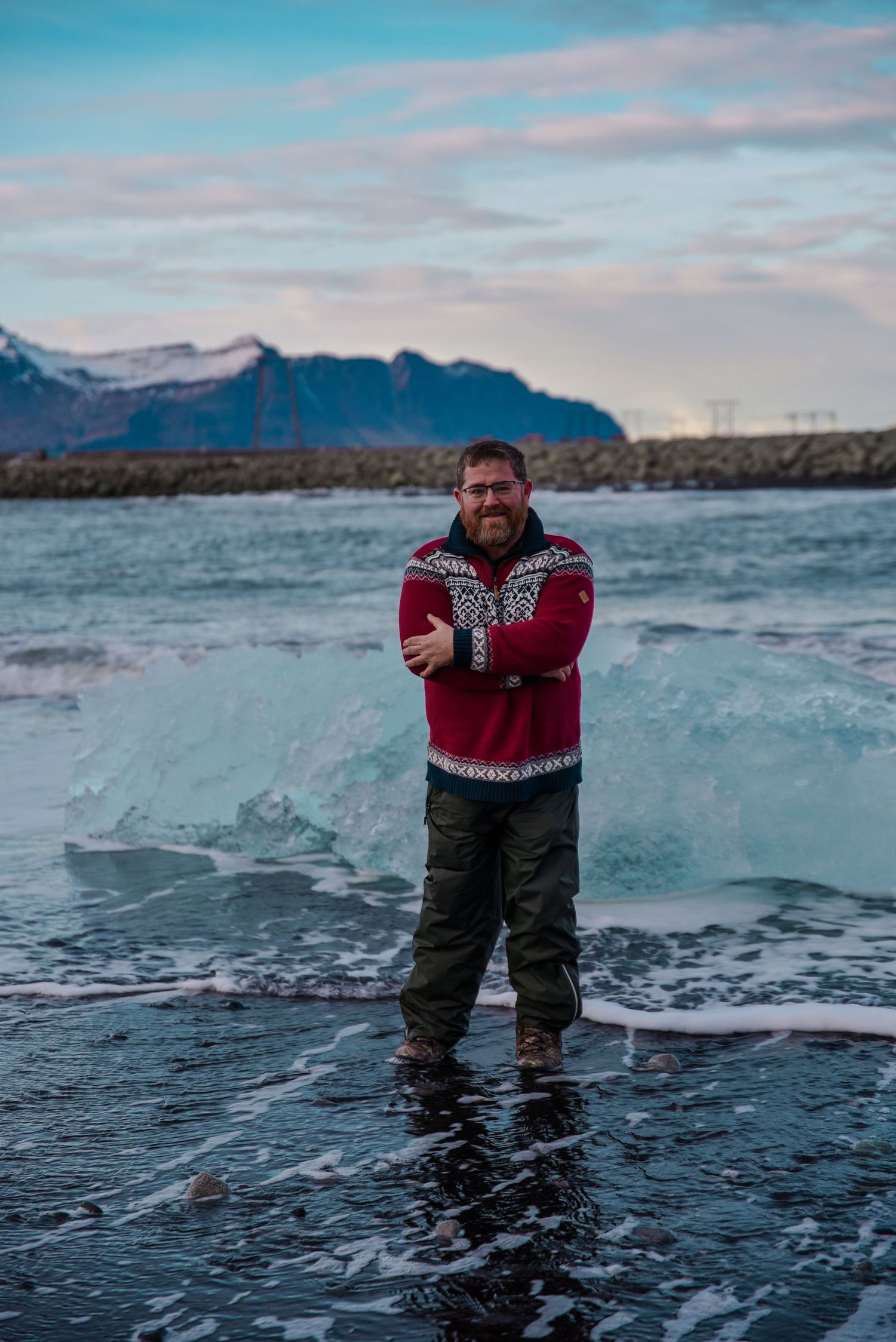 Bearded man in red Nordic sweater standing on icy glacier beach in Iceland with snow-capped mountains behind.