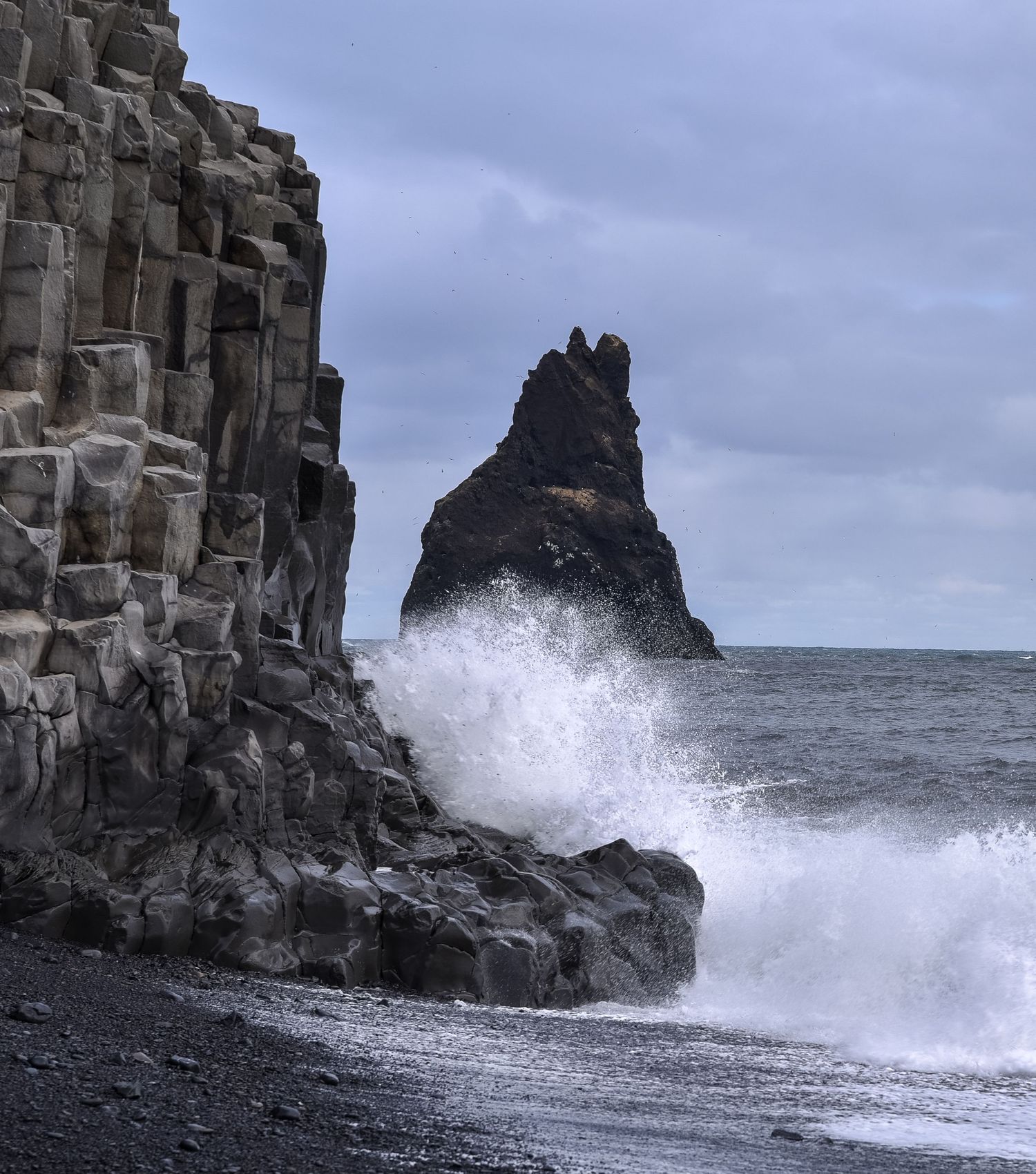 Massive waves crash against black basalt columns on a volcanic beach with a sea stack in the distance.