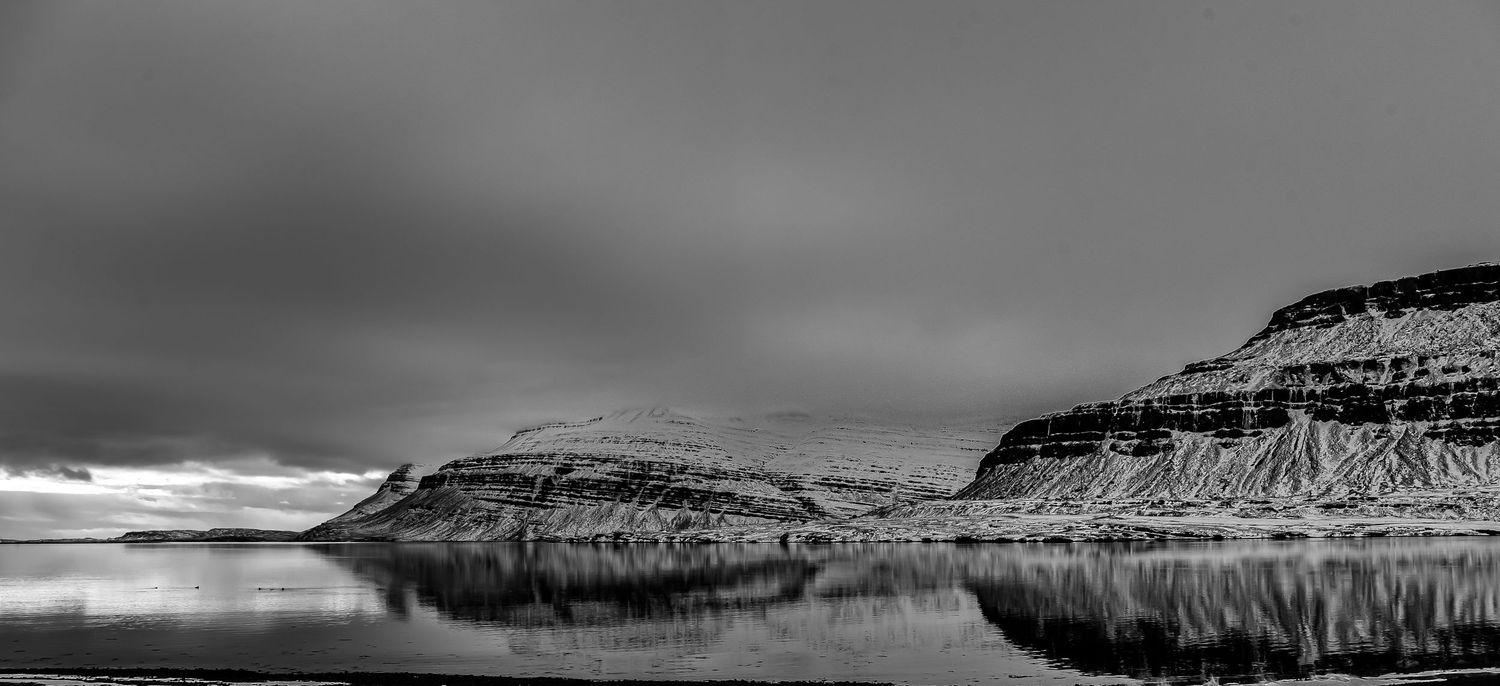 Black and white panoramic view of a calm fjord reflecting snow-capped cliffs in Iceland.