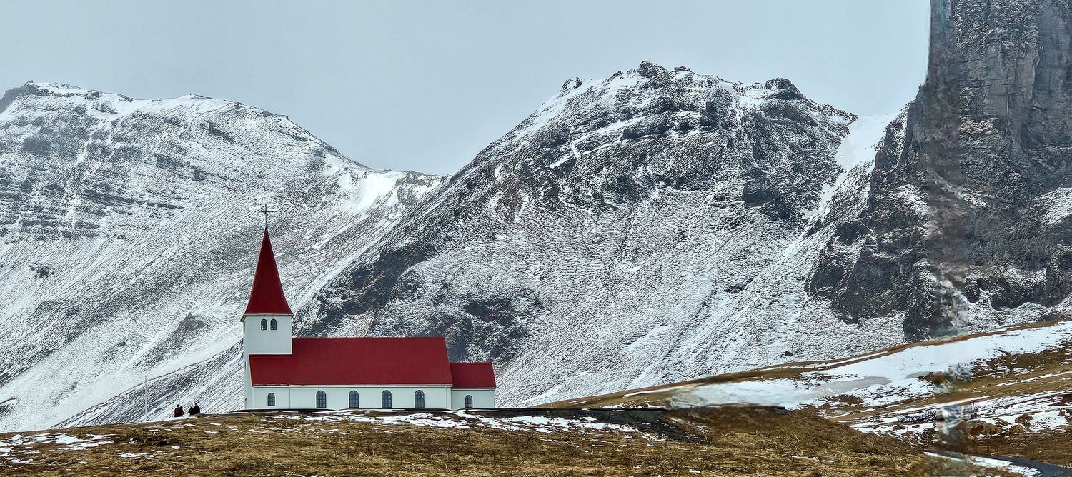 Small white church with red roof nestled below dramatic snow-covered mountains in Iceland.