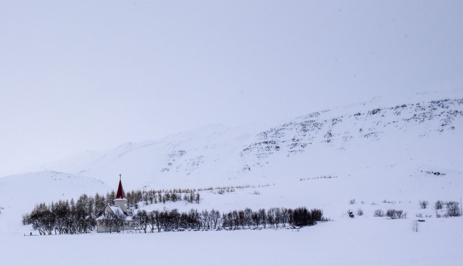 A red-steepled church surrounded by bare trees in a vast snow-covered Icelandic winter landscape.