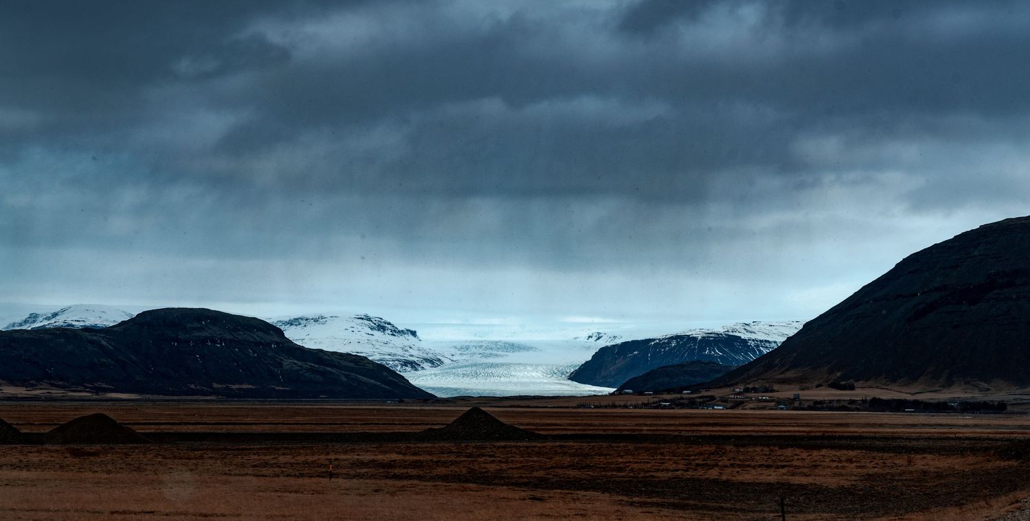 Dramatic stormy sky looms over a vast Icelandic plain with snow-capped mountains and a distant glacier.