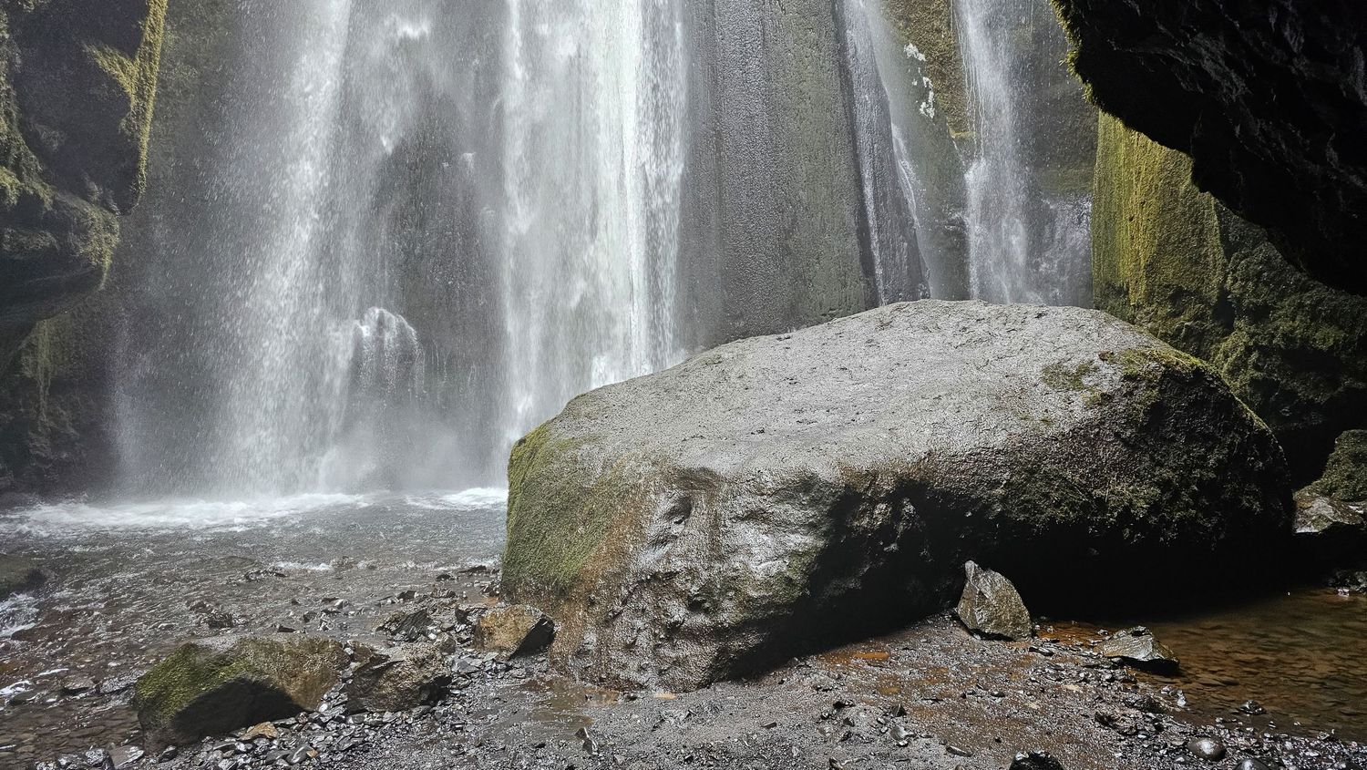 Powerful waterfall cascades behind a large mossy boulder in a rocky grotto.
