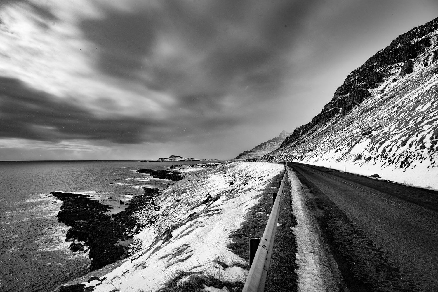Black and white coastal road stretches along rocky shoreline beneath dramatic cloudy winter sky.