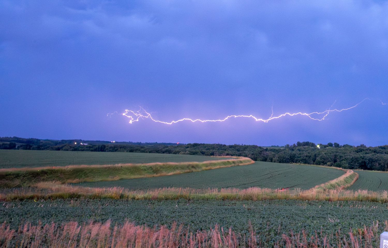 Horizontal lightning bolt stretching across a purple stormy sky over green farmland fields.
