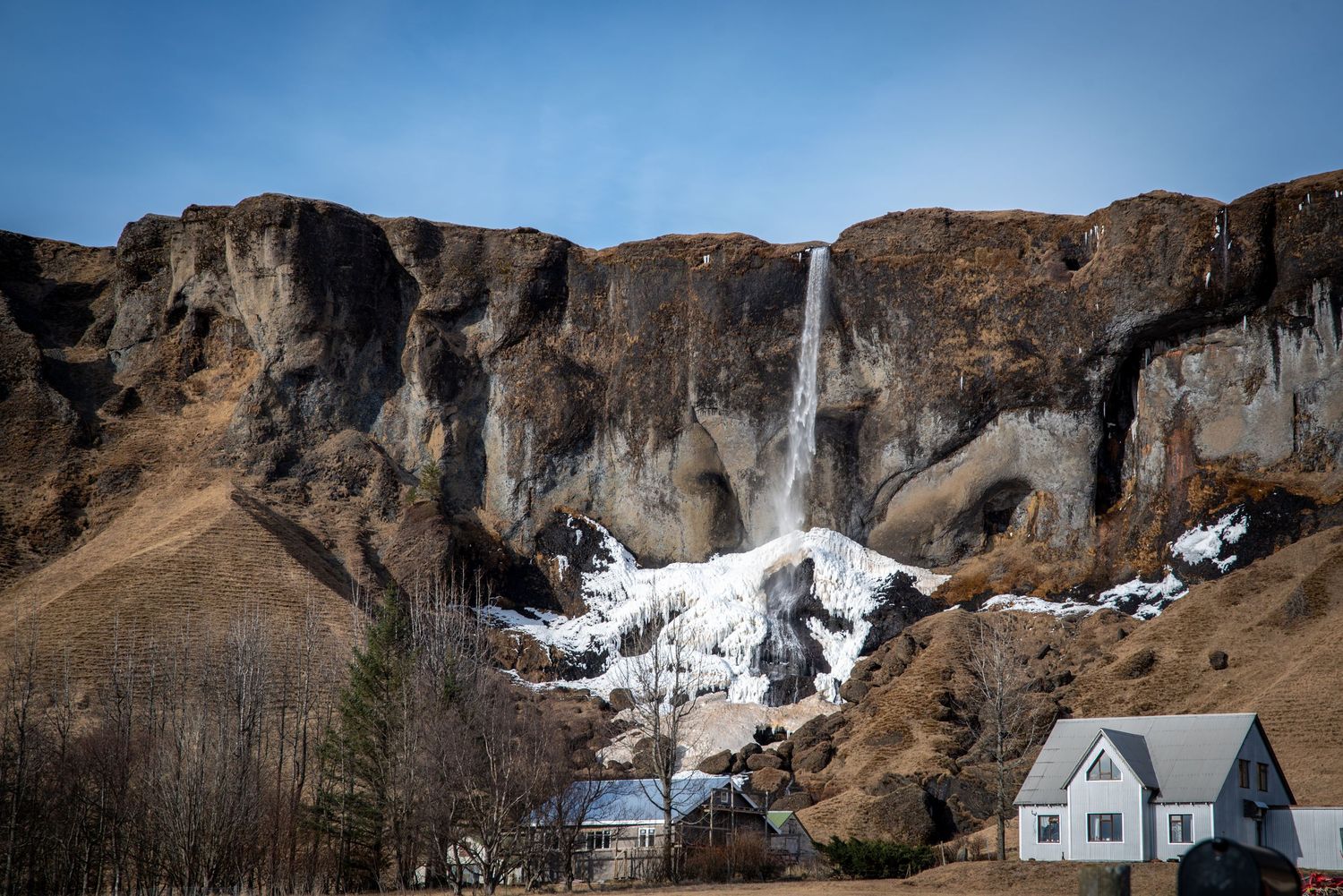Dramatic waterfall cascading down rocky cliffs with snow beside a white house in an Icelandic valley.
