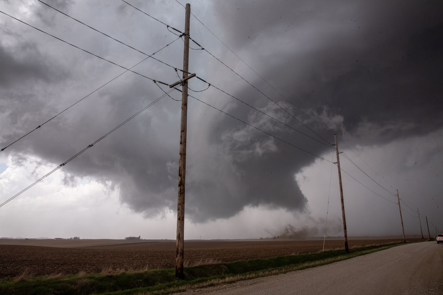 Massive tornado forming over flat farmland with dark storm clouds and power lines in foreground.