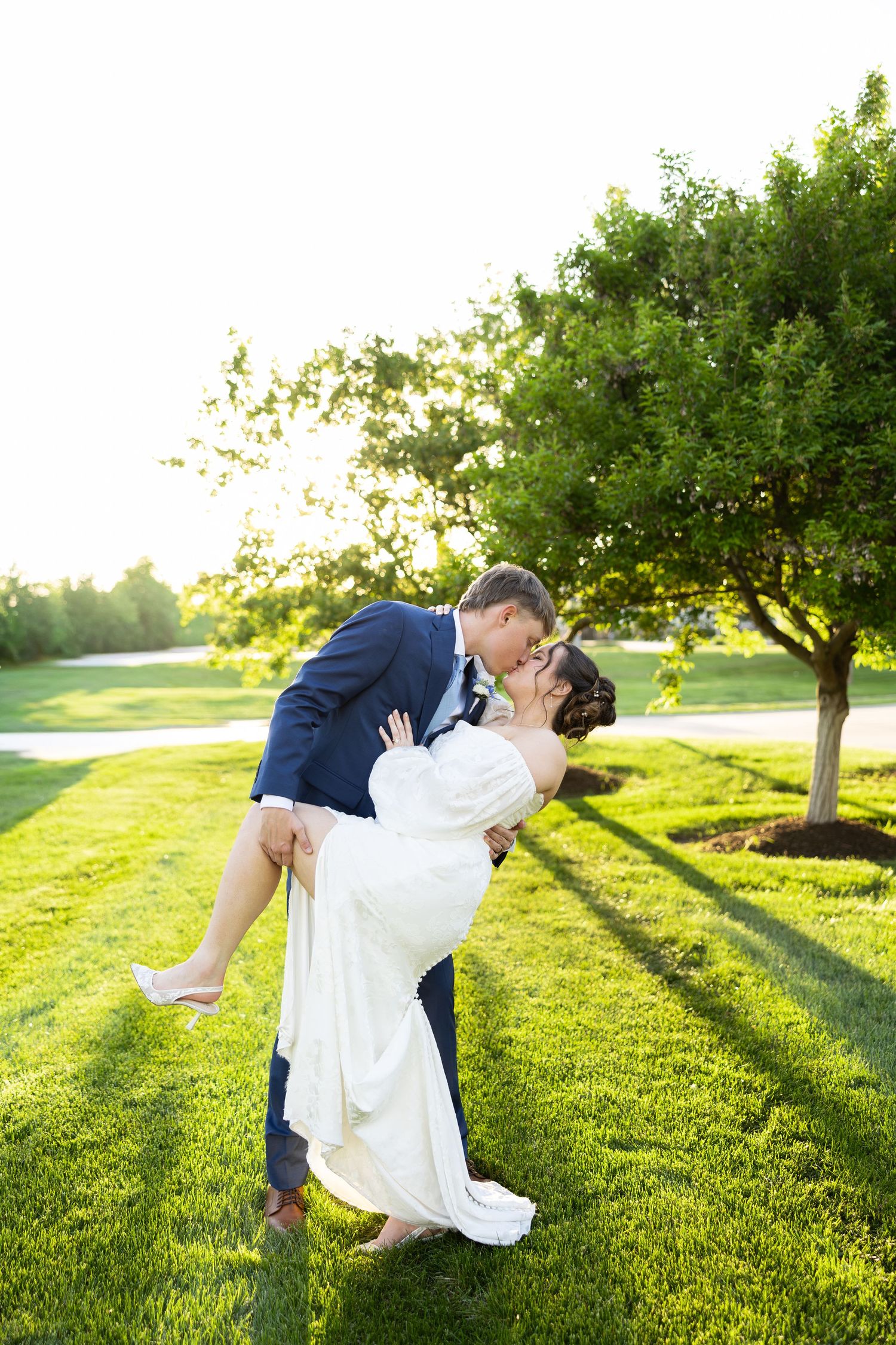 Groom in navy suit dips bride in white gown for a romantic kiss on lush green lawn at golden hour.