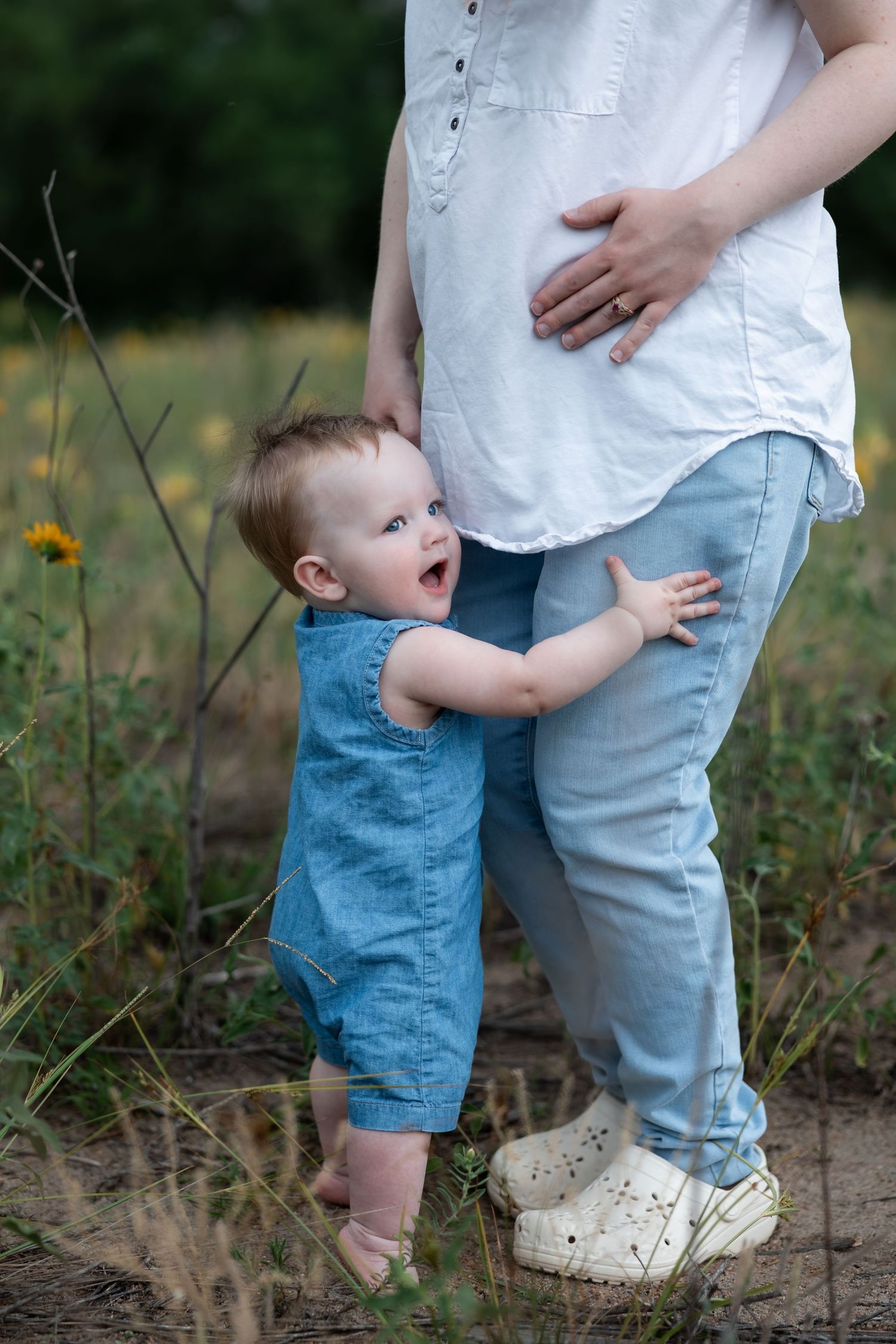 Toddler in denim romper hugging pregnant mother's legs outdoors in a field with wildflowers.